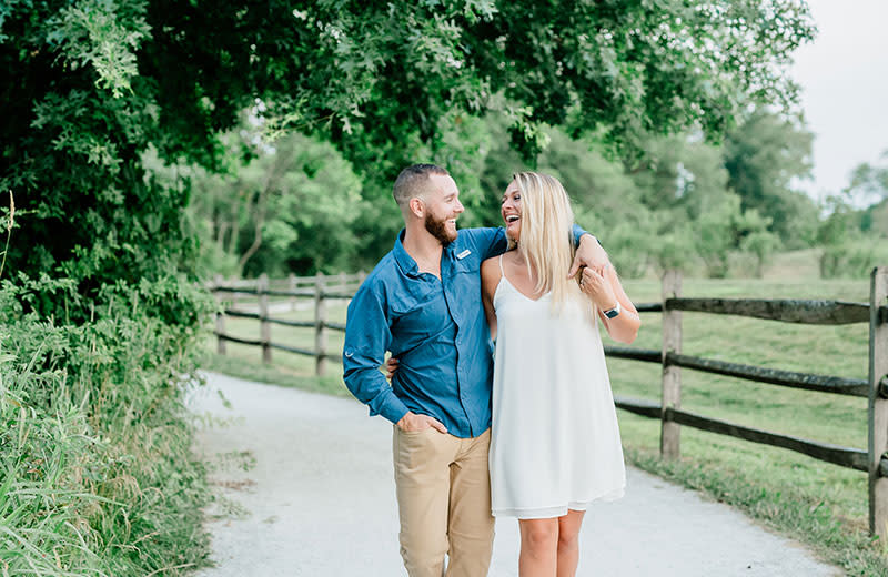 A young married couple share an embrace while walking on a paved path next to a forest.