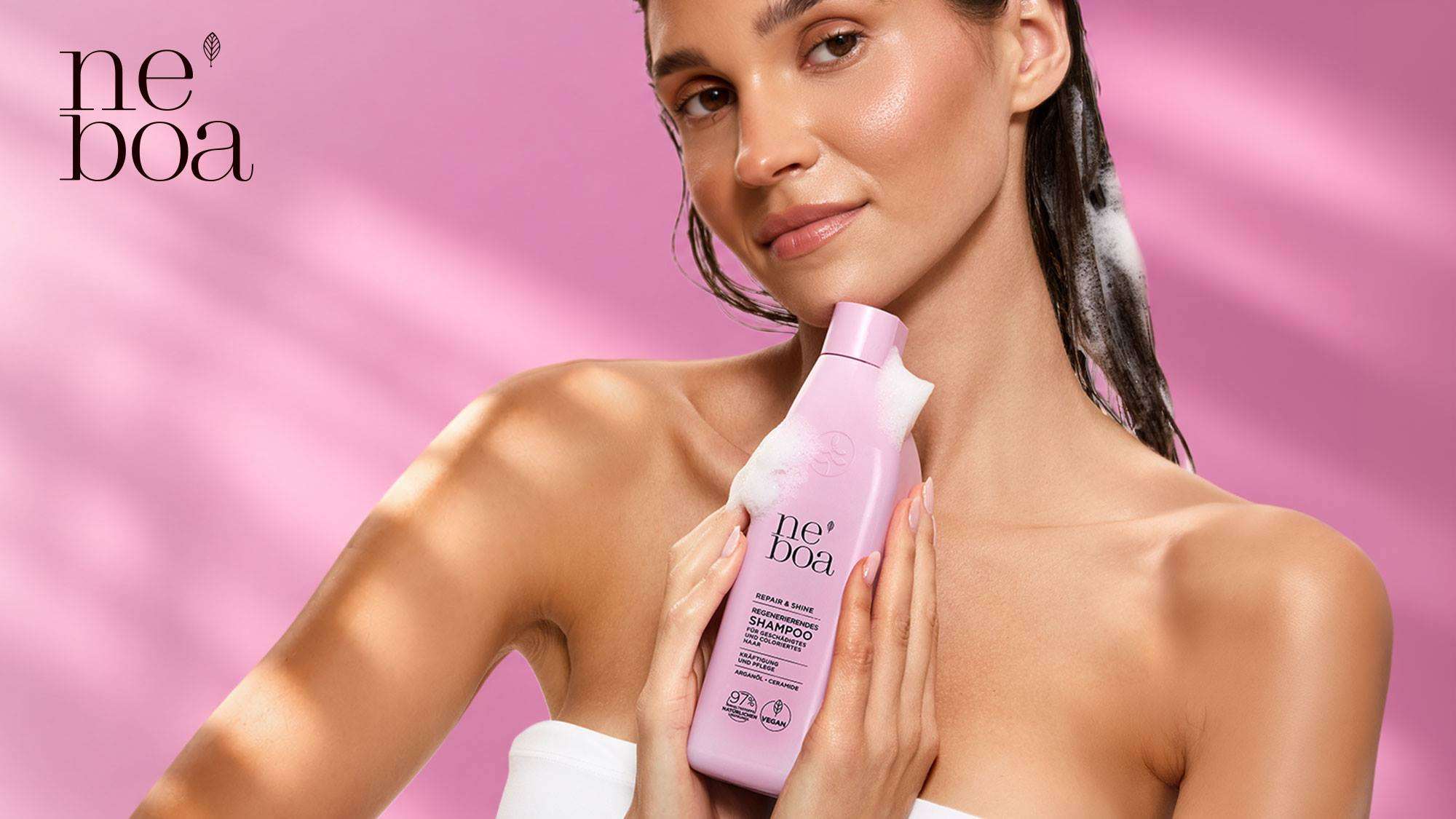 Werbemotiv der Marke Neboa: Eine Frau mit feuchtem Haar hält eine rosa Flasche Neboa Repair & Shine Shampoo vor rosa Hintergrund. Aufschrift der Flasche: Neboa Repair & Shine Regenerierendes Shampoo für geschädigtes und coloriertes Haar, Kräftigung und Pflege mit Arganöl und Ceramiden, 97 Prozent Inhaltsstoffe natürlichen Ursprungs, vegan. Verlinkung zu den Produkten