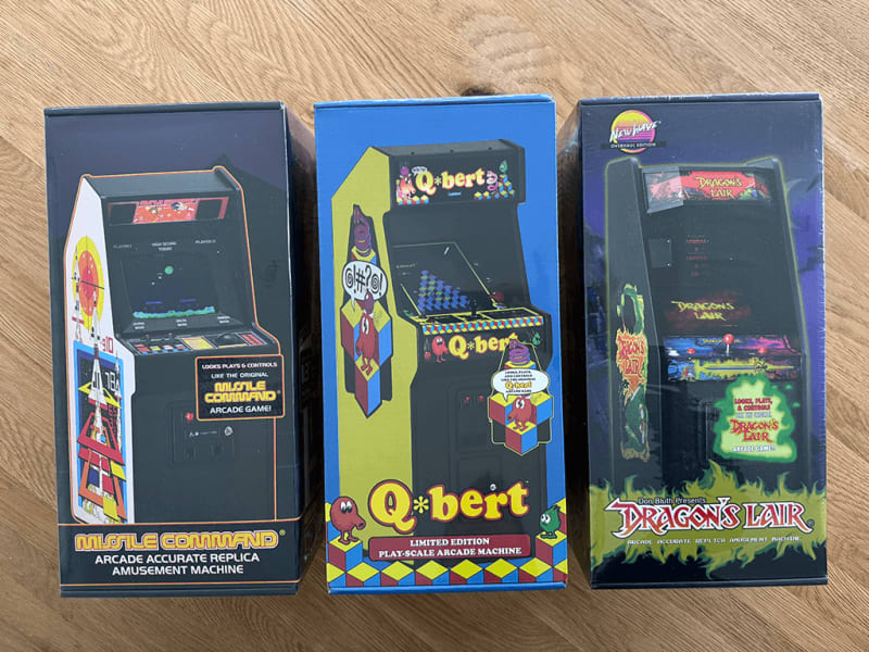 Boxed arcades Three boxed New Wave 1/6 scaled arcade cabinets, Missle Command, Qbert and Dragons Liar on a wooden table top surface.