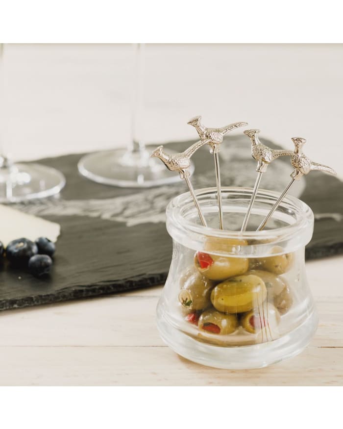 a glass jar with olives and a couple of glasses on a table