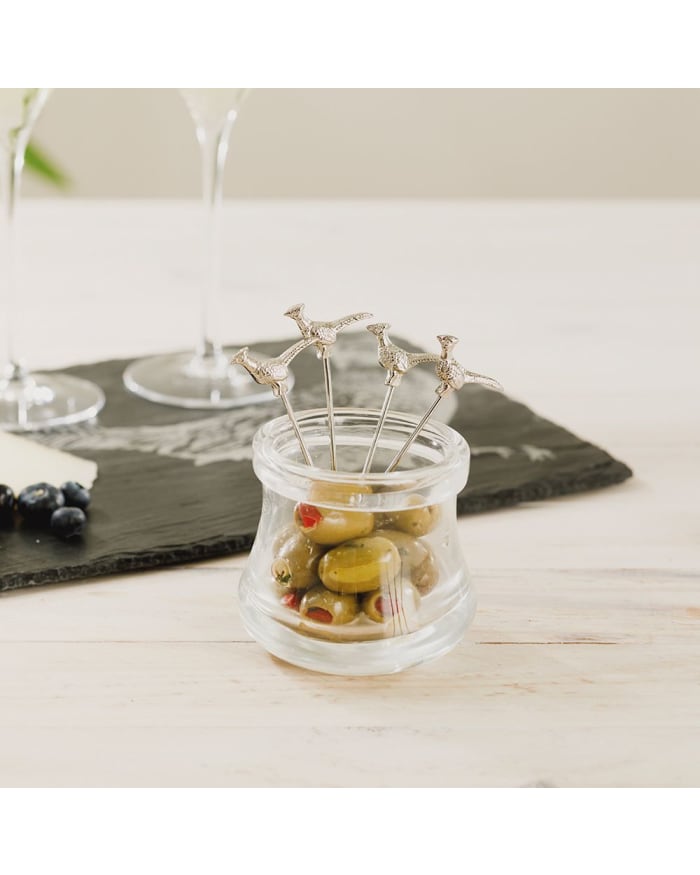 a glass jar with olives and a couple of glasses on a table