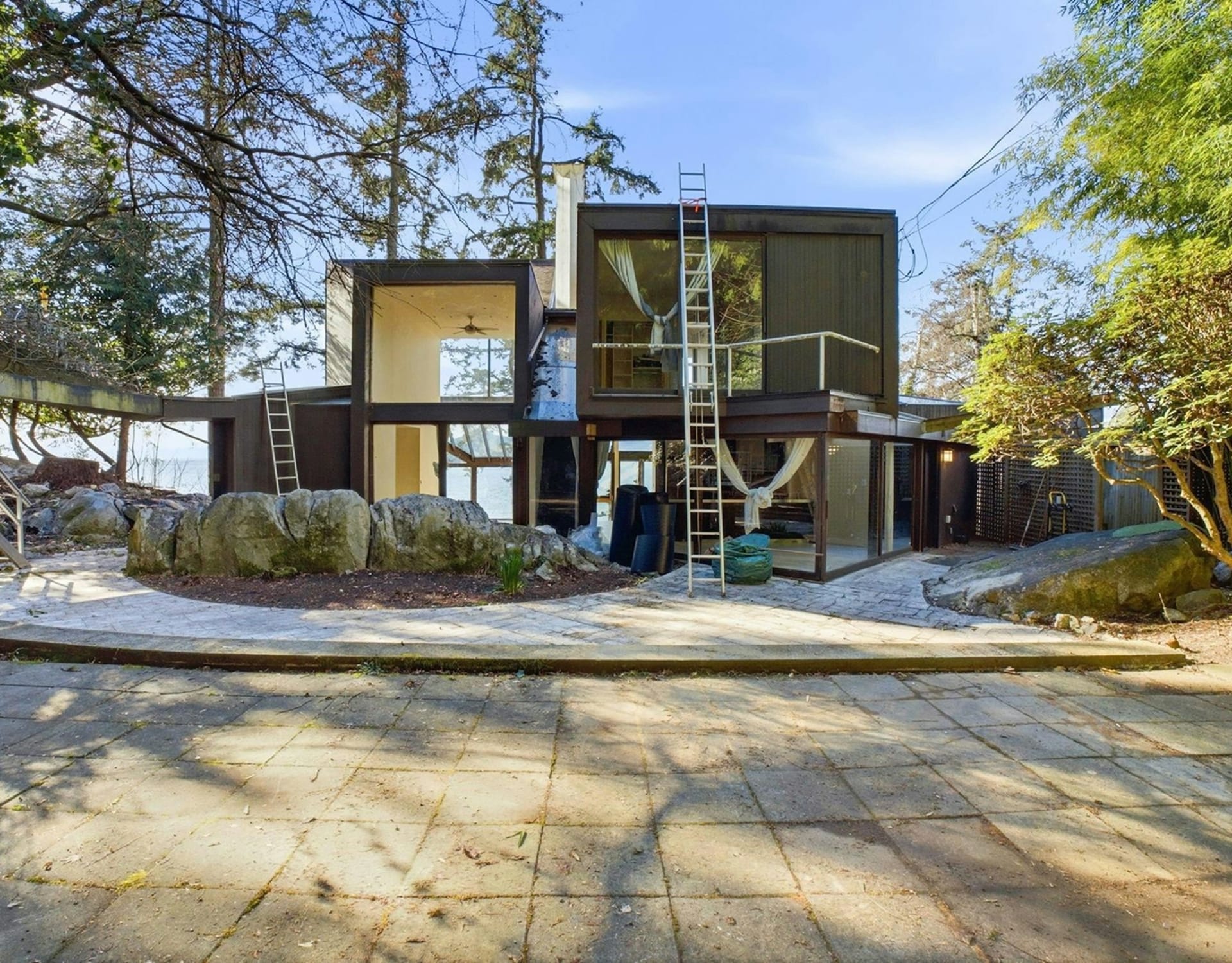 A modern, dark-paneled house with large windows sits on a rocky shore, with a paved patio in the foreground and lush trees in the background.