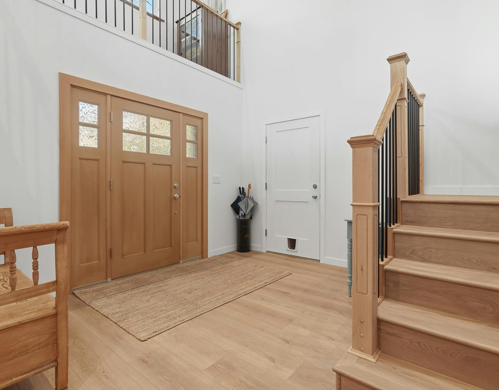 A light wood entryway with a glass-paneled double door and a jute rug in the foreground, and a staircase and loft railing in the background.