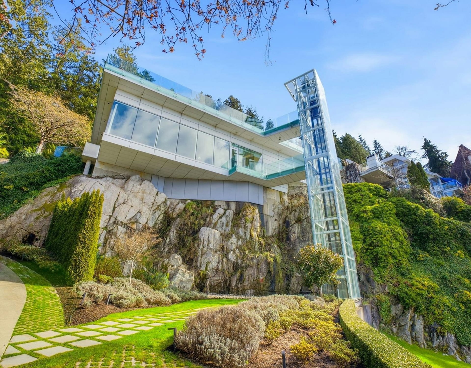 A modern glass elevator ascends a rocky cliff face towards a sleek white house with large windows, set against a backdrop of lush green hills and trees under a clear blue sky.