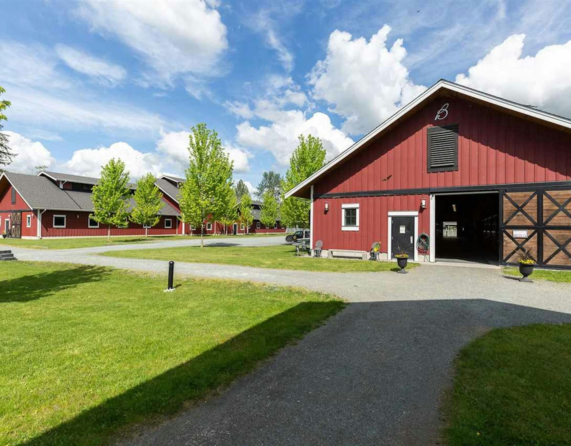 A paved path leads through a green lawn toward several red barns under a bright, cloudy blue sky.