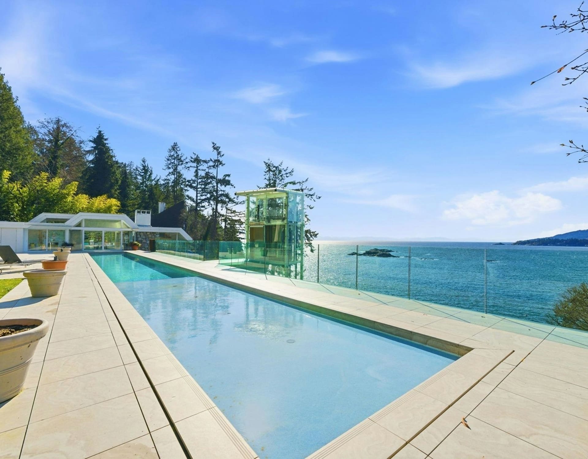 A long, rectangular swimming pool with a modern glass elevator sits in the foreground, overlooking a vast blue ocean and distant islands under a clear sky.