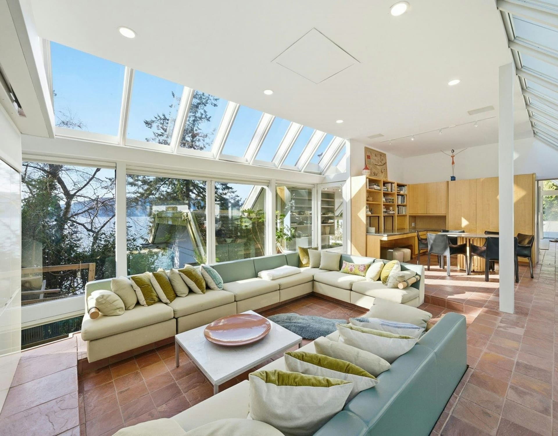 A large sectional sofa with many pillows sits on a terracotta tiled floor in a sunlit room with a glass ceiling and walls overlooking a body of water and trees, while a wooden built-in bookshelf and dining area are visible in the background.