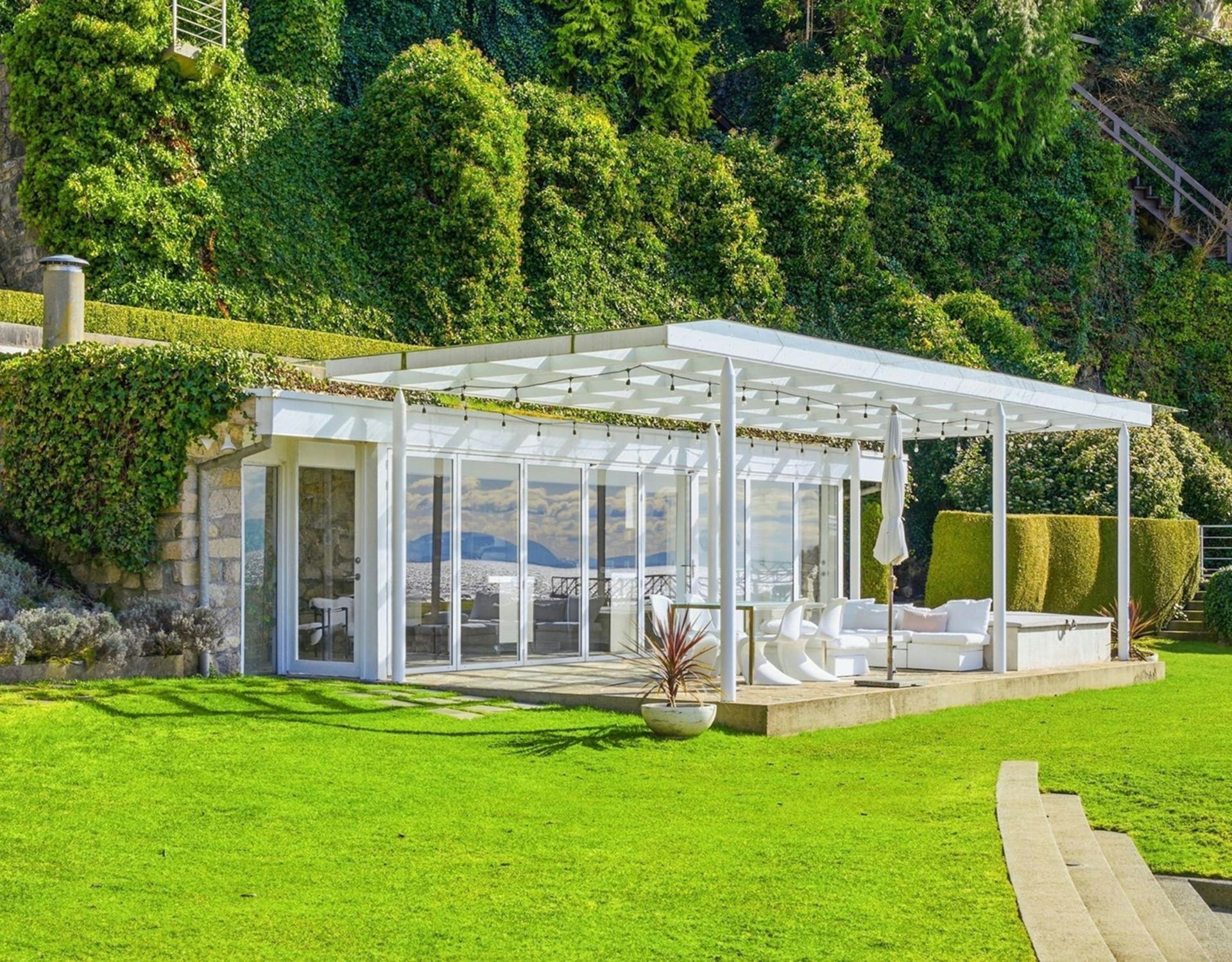 A modern white pavilion with glass walls and outdoor seating sits on a lush green lawn, backed by a steep, tree-covered hillside.