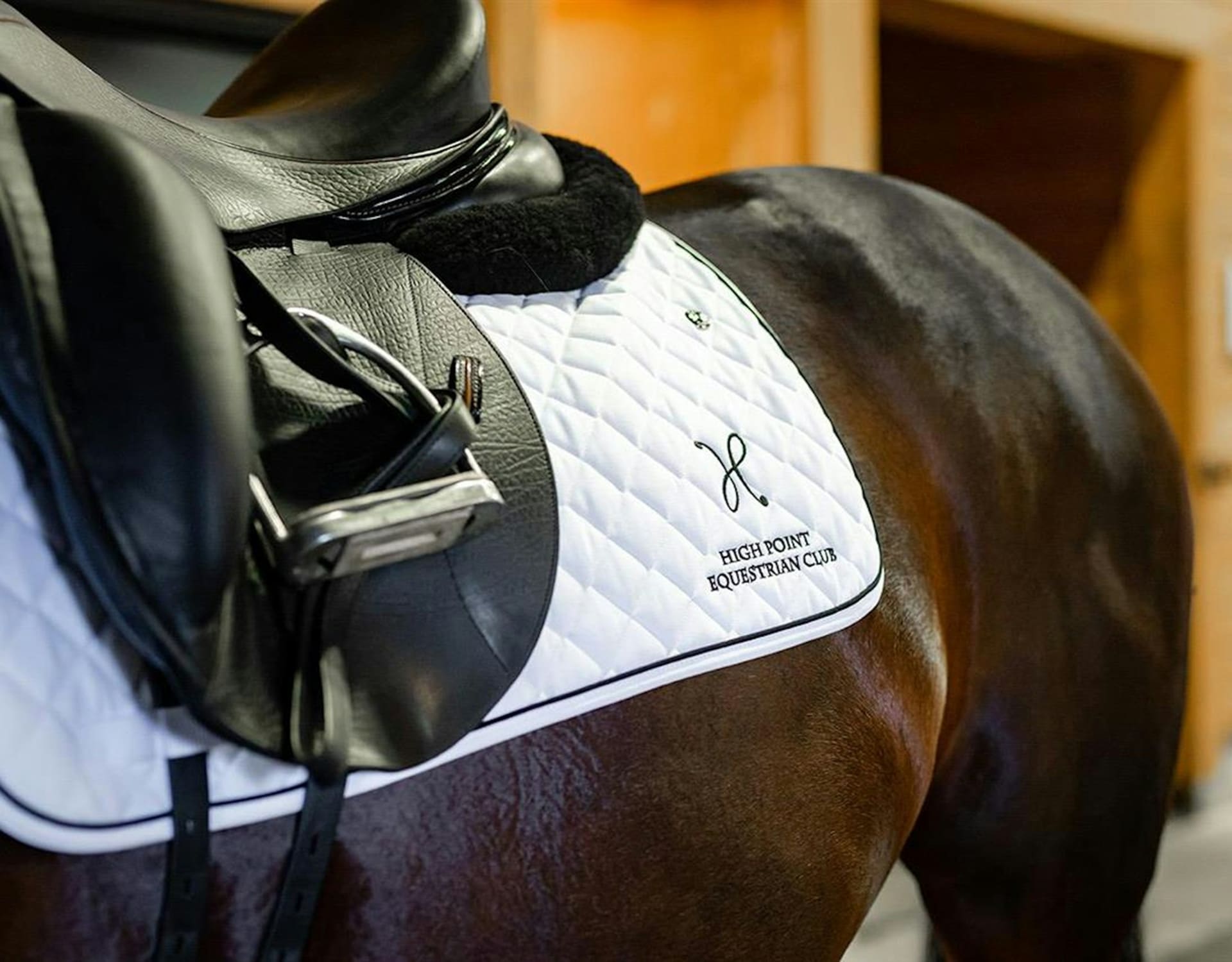 A black leather saddle rests on a white quilted saddle pad atop a brown horse, set against a blurred wooden stable background.
