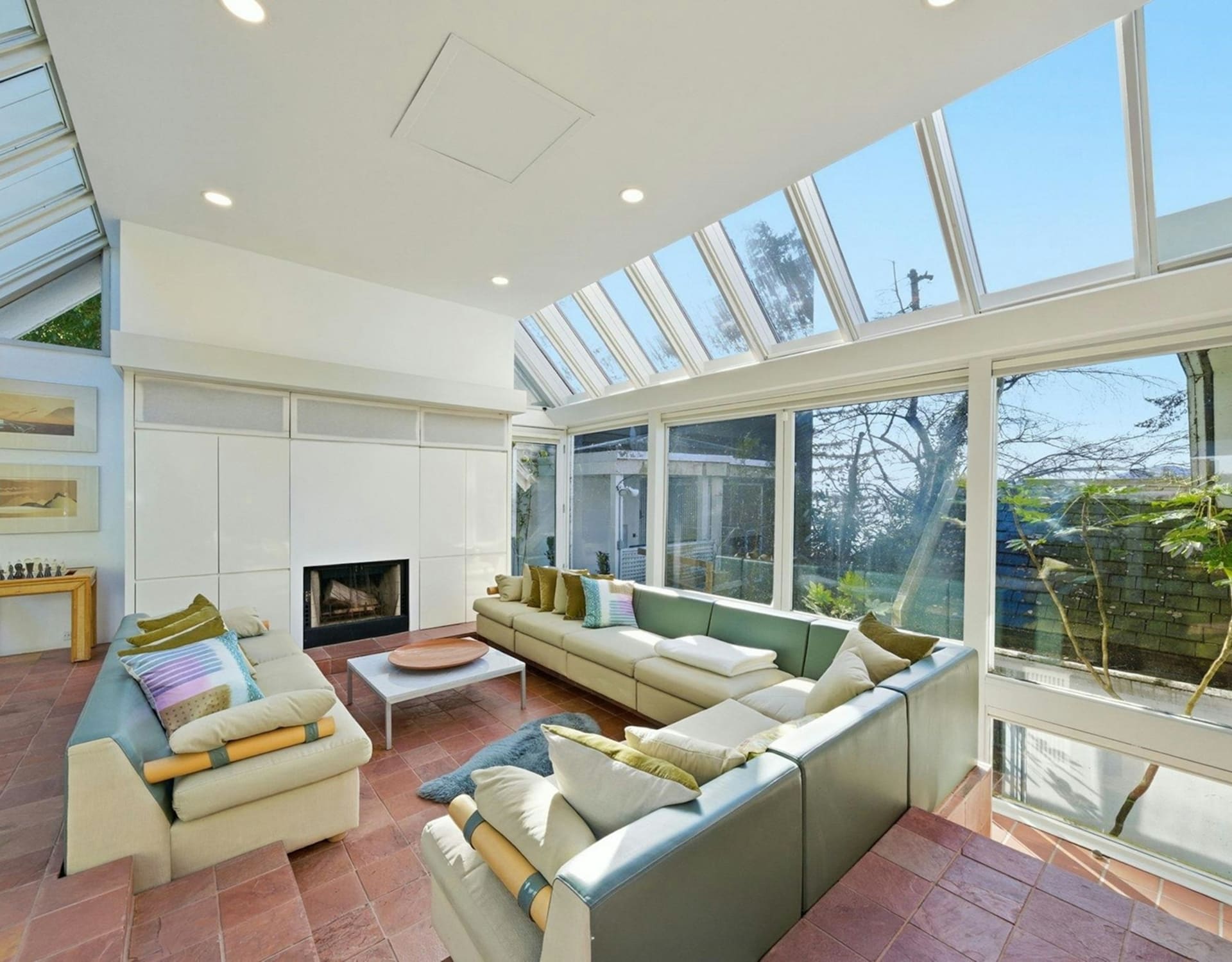 A spacious living room with a large sectional sofa and a fireplace in the foreground is bathed in natural light from a glass ceiling and large windows that overlook a lush green backyard.