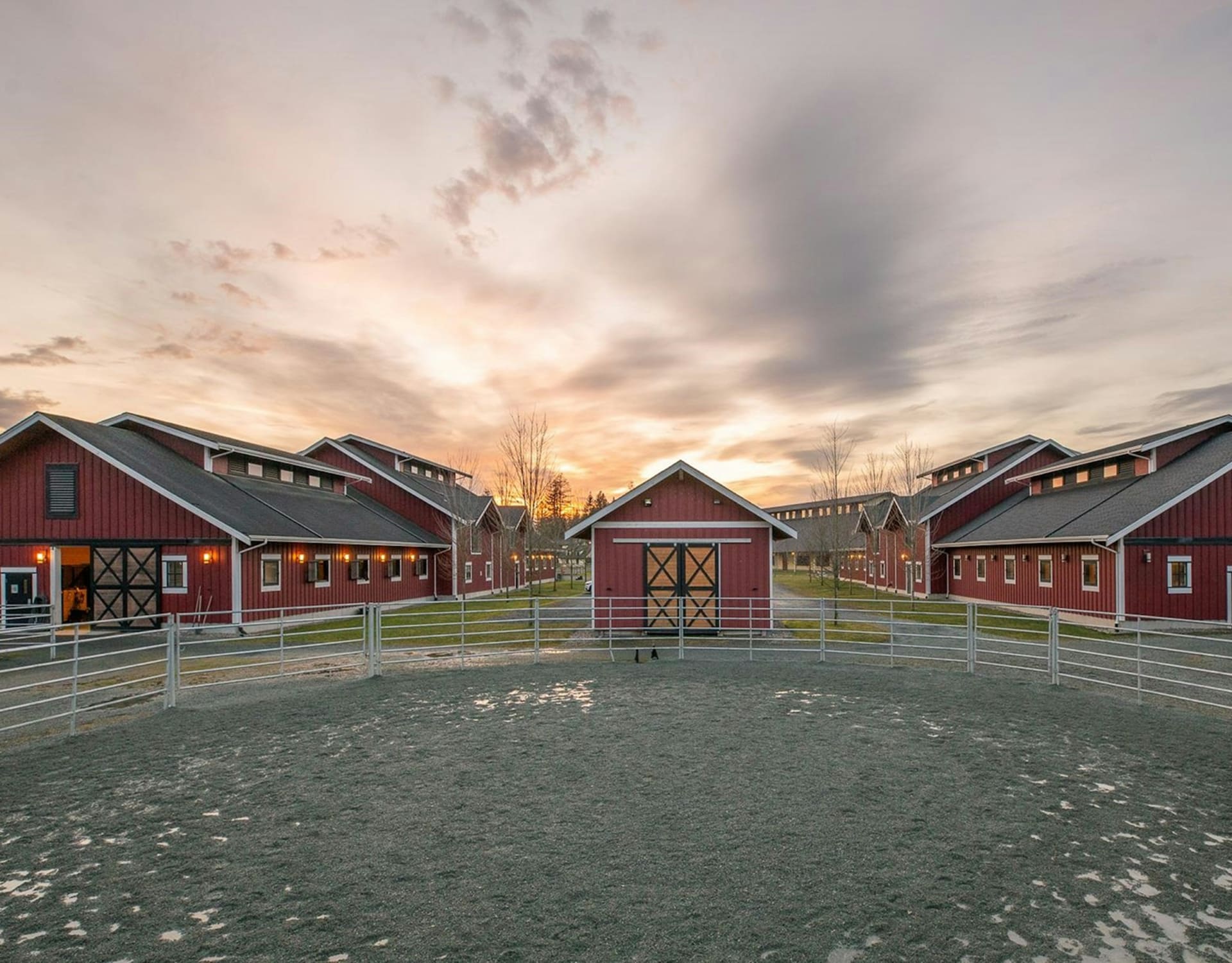 A sandy paddock is enclosed by metal fencing in the foreground, surrounded by red barns under a dramatic sunset sky.