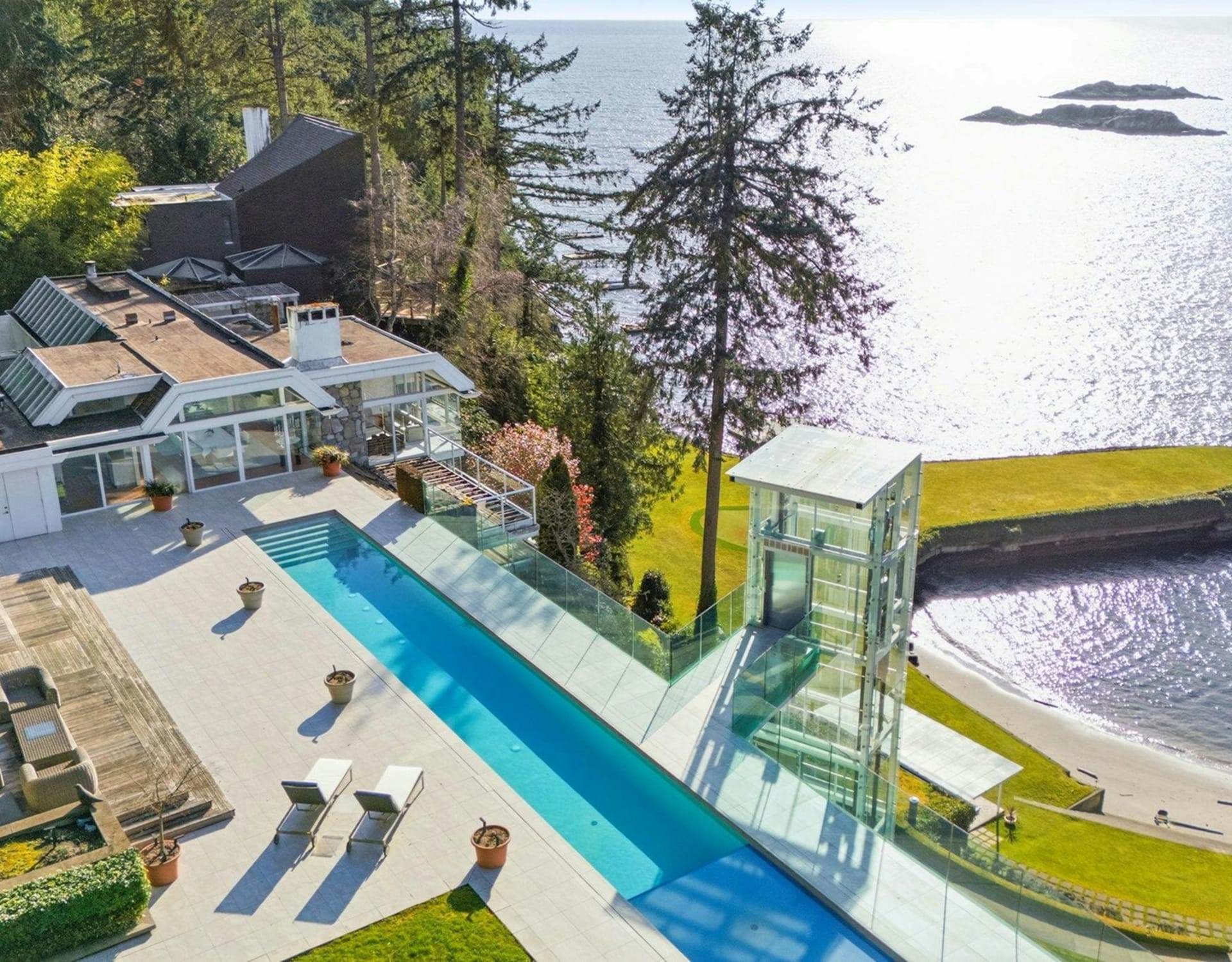 A modern mansion with a long swimming pool and glass elevator sits on a cliff overlooking a sparkling ocean with small islands in the distance.