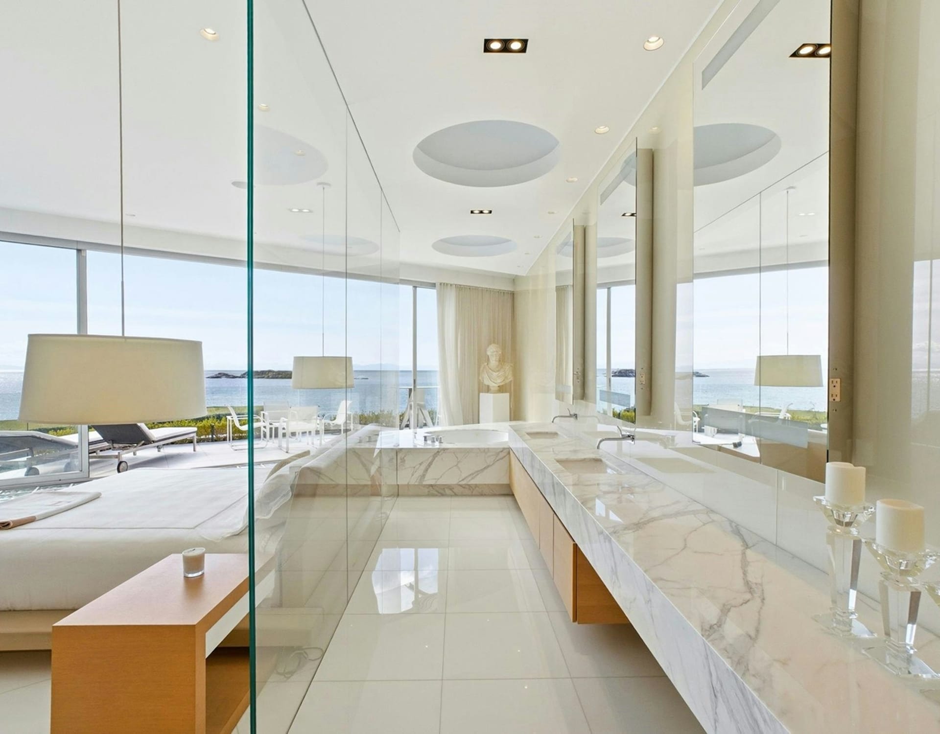 A modern bathroom with a marble vanity and large mirrors is in the foreground, while a bedroom with a bed and a view of the ocean and coastline is in the background.