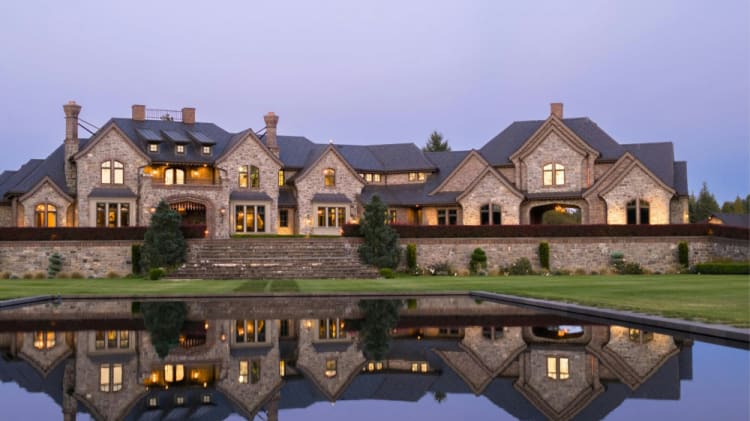 A large, luxurious stone and wood mansion with multiple chimneys and gabled roofs is reflected in a calm, still pool of water, creating a symmetrical and serene scene against a twilight sky.