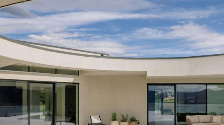 A modern patio with minimalist furniture sits before a curved building with large glass doors, all under a bright blue sky with scattered clouds.