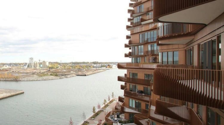 A modern building with curved balconies overlooks a calm waterway and industrial background.