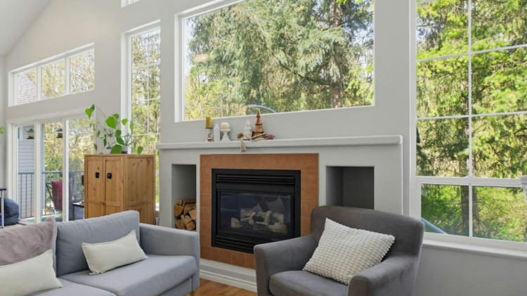 A cozy living room with large windows overlooking a lush, forested landscape, featuring a fireplace, comfortable gray sofas, and decorative accents.