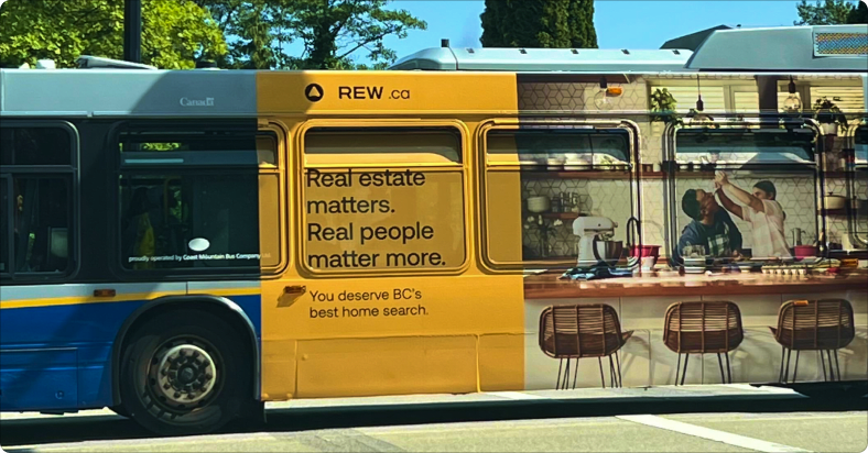 A TransLink bus showing on REW ad on the side of it depicting a father and child in a kitchen with the words "Real estate matters. Real people matter more" next to it.