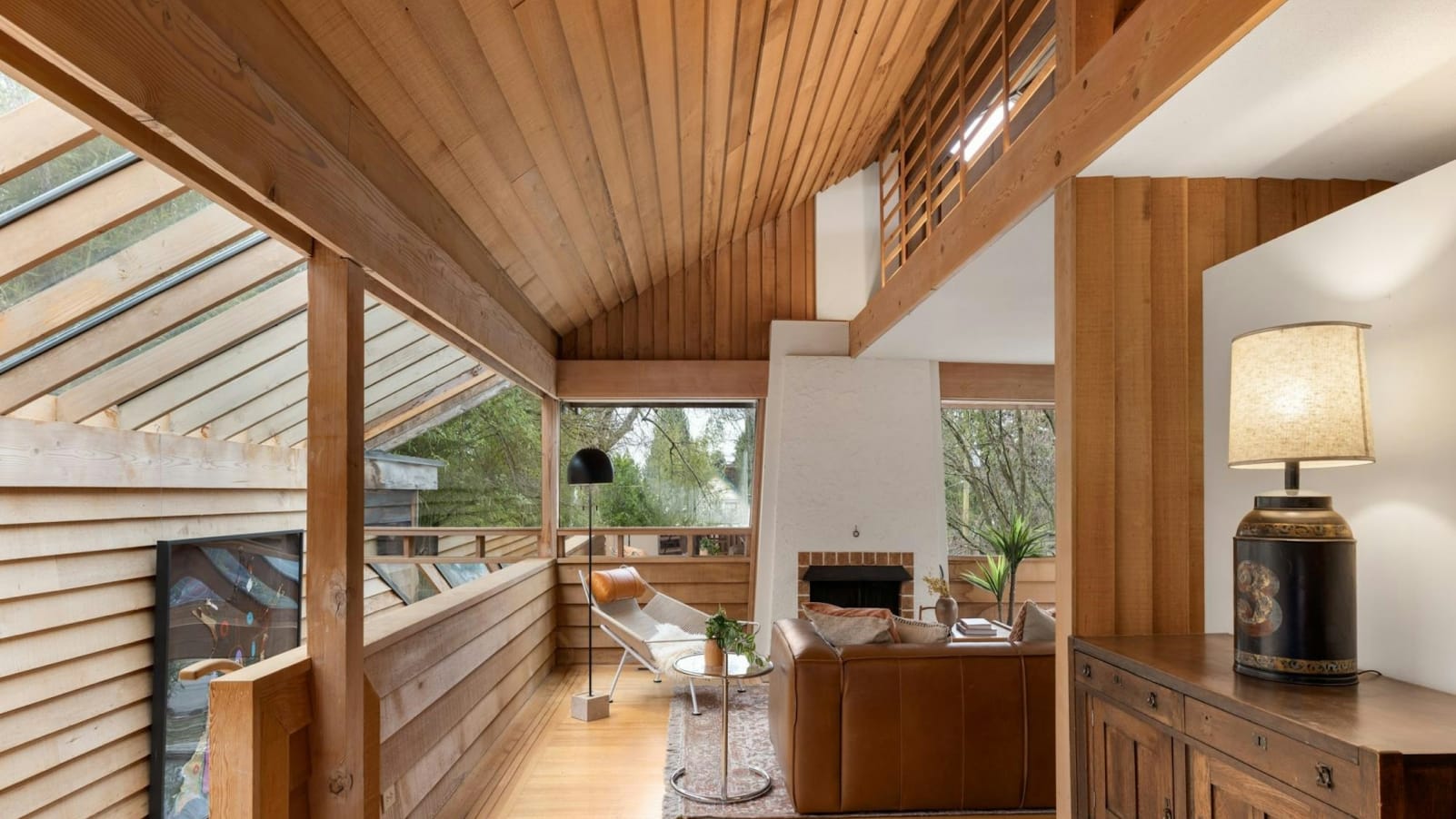 A cozy and inviting interior space with wooden paneled walls, a fireplace, and large windows overlooking a natural outdoor setting.