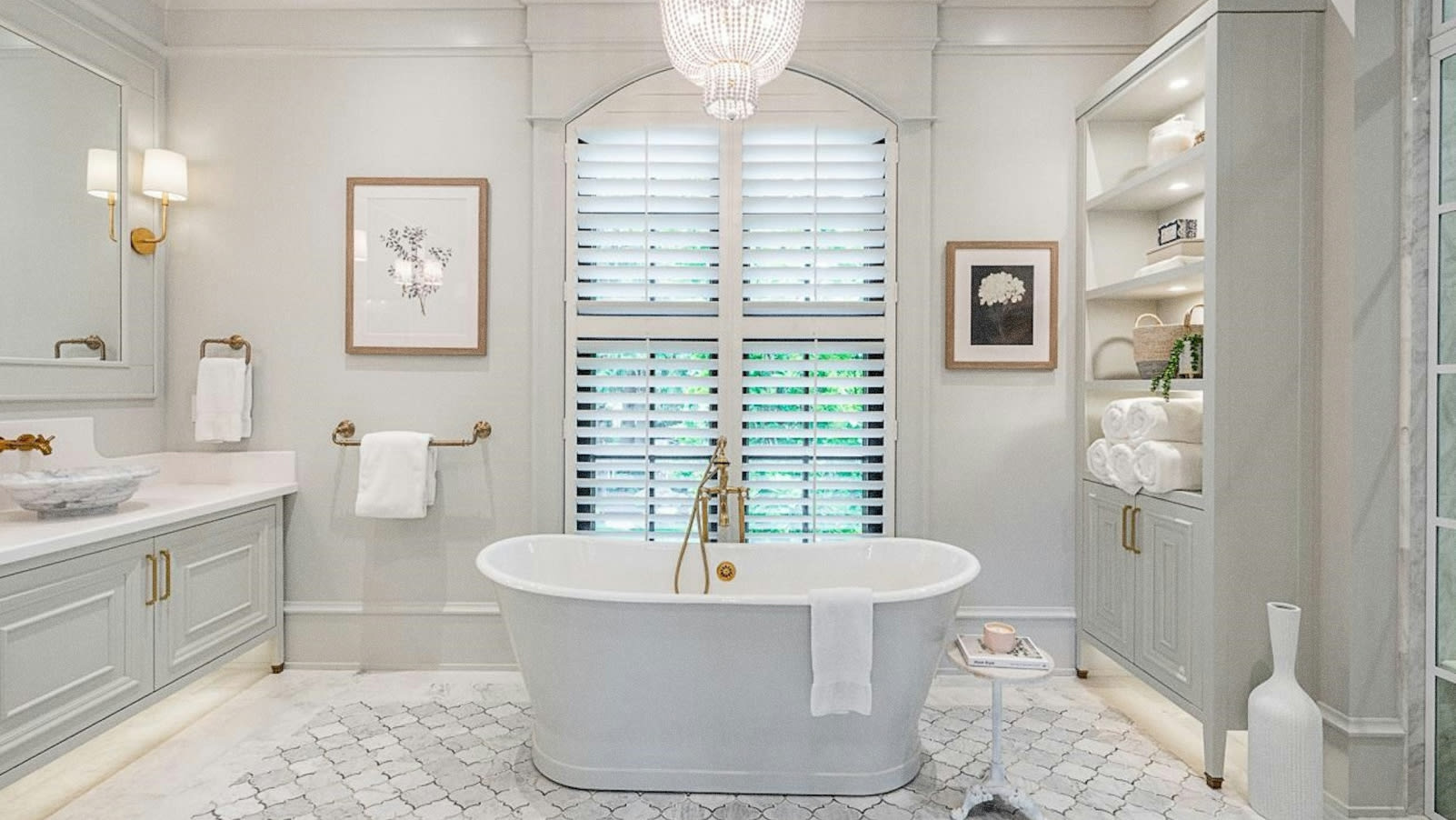 A spacious and elegantly designed bathroom with a freestanding bathtub, white shuttered windows, and various decorative elements such as framed artwork and shelving.