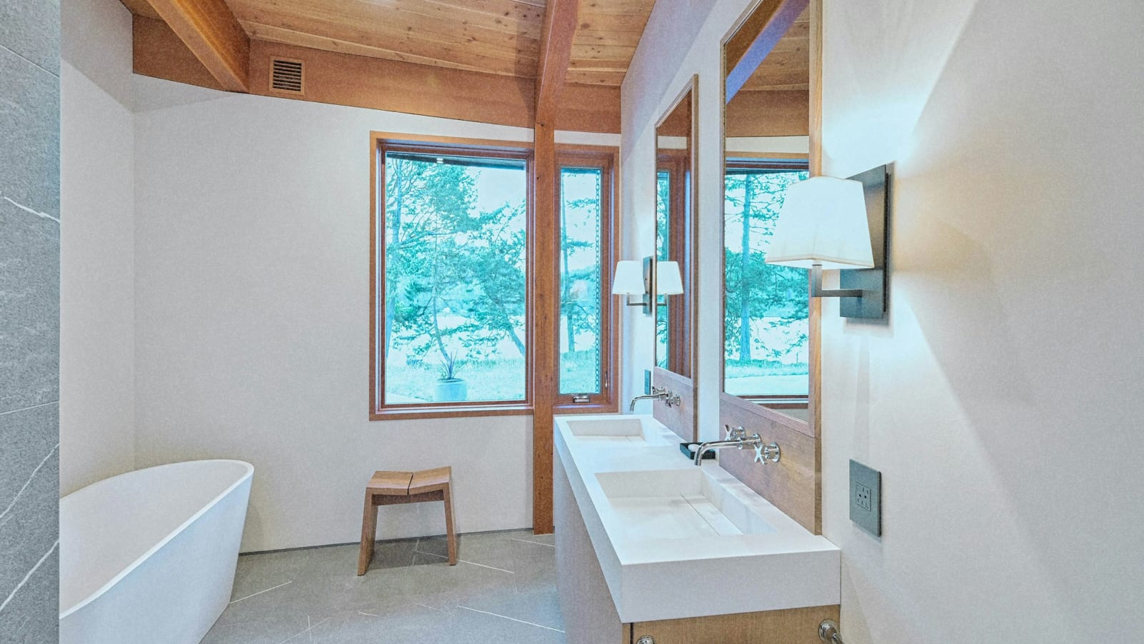 A modern bathroom with a large window overlooking a lush, green outdoor environment, featuring a white bathtub, a wooden stool, and wall-mounted lighting fixtures.