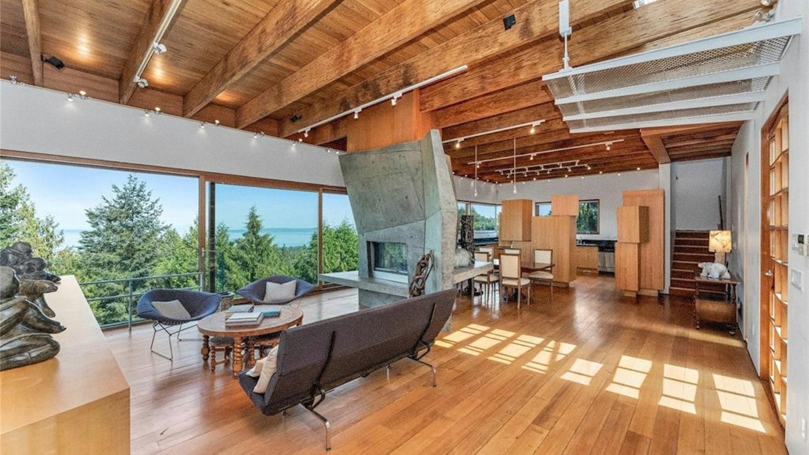 A spacious and modern living room with wooden beams, large windows overlooking a forested landscape, and a mix of contemporary and rustic furnishings.