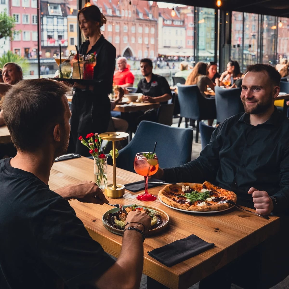 Goście przy stole z pizzą w sali Riverside z widokiem na kamienice nad Motławą