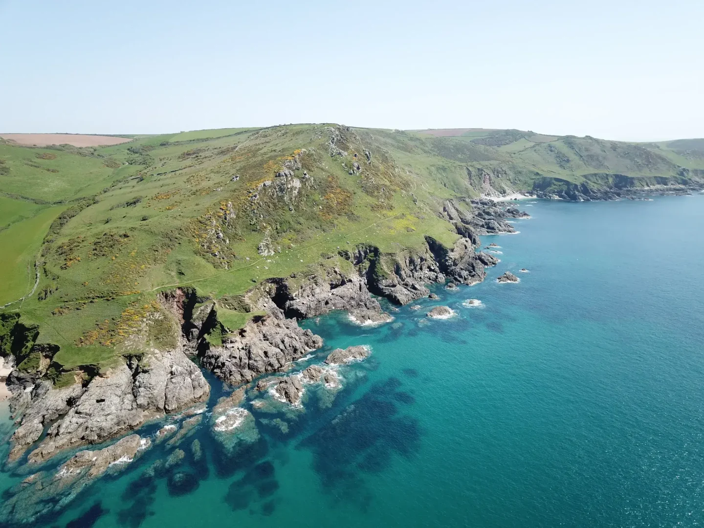 Aerial view of the Devon coastline with rugged cliffs, green rolling hills and a clear turquoise sea under a bright blue sky.