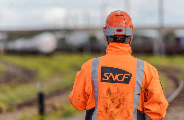 Employés vu de dos avec logo SNCF sur la veste orange avec train en flou à l'arrière plan