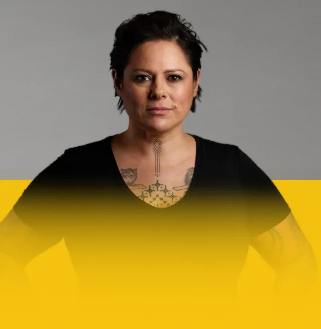 Anika Moa wears a black top with hands on hips, looking straight ahead to camera, on a light grey and yellow background.