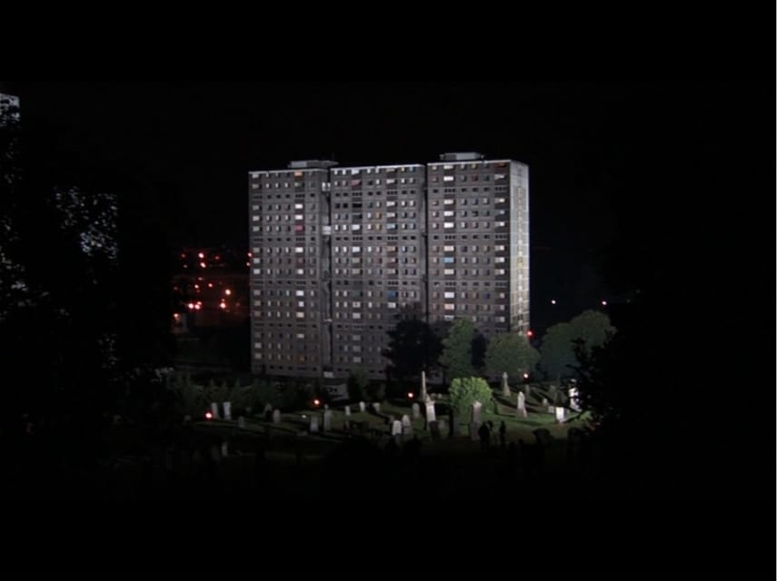A grey concrete residential tower block of at least 18 floors in Glasgow is pictured at night. Illuminated in front of the imposing building are large trees and what look like gravestones.