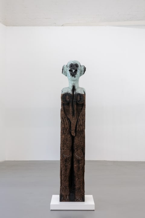 Carved styrofoam and cork sculpture, stood on a very low white plinth. The sculpture has a brown body with gender non-specific features carved in, a greenish head with very large ears and dark cavities for eyes.