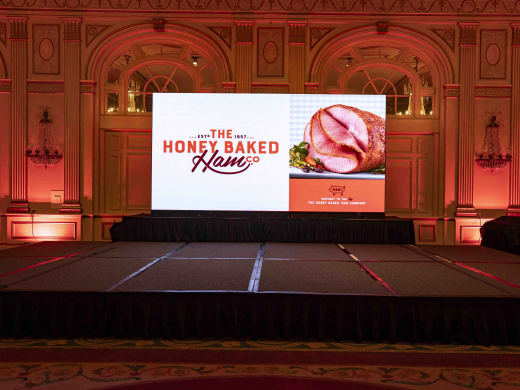 HoneyBaked Ham brand-reveal LED screen with the 'Est 1957' logo against a red-washed historic ballroom