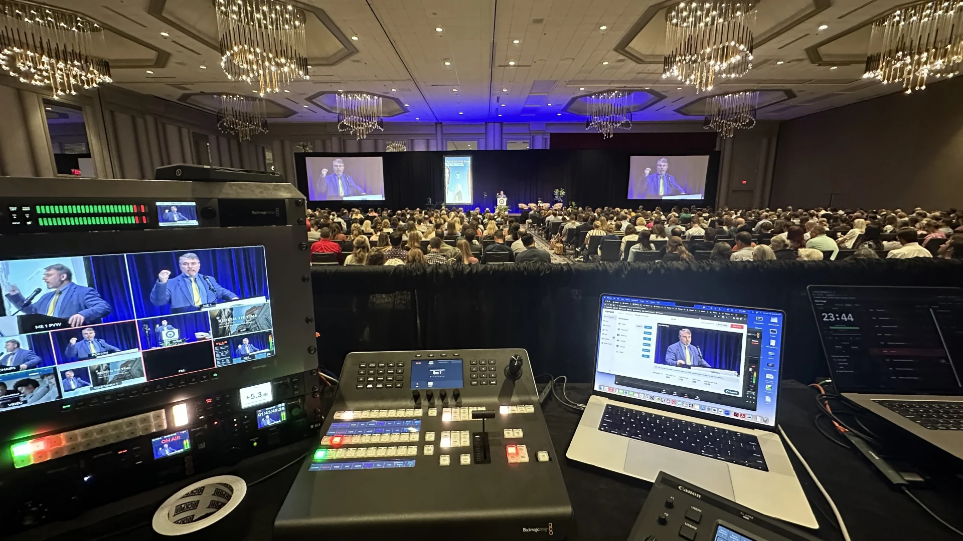 Conference video control desk with multiviewer, switcher, and streaming laptop facing a packed general session with IMAG screens