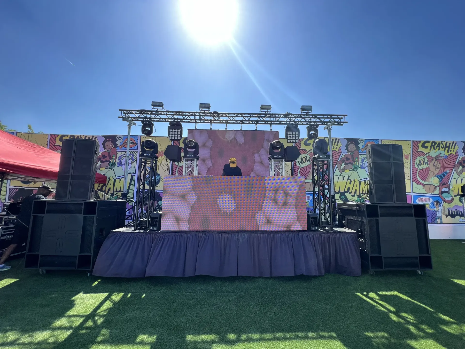DJ performing on deck stage with LED wall, truss lighting, and subwoofers against pop art backdrop