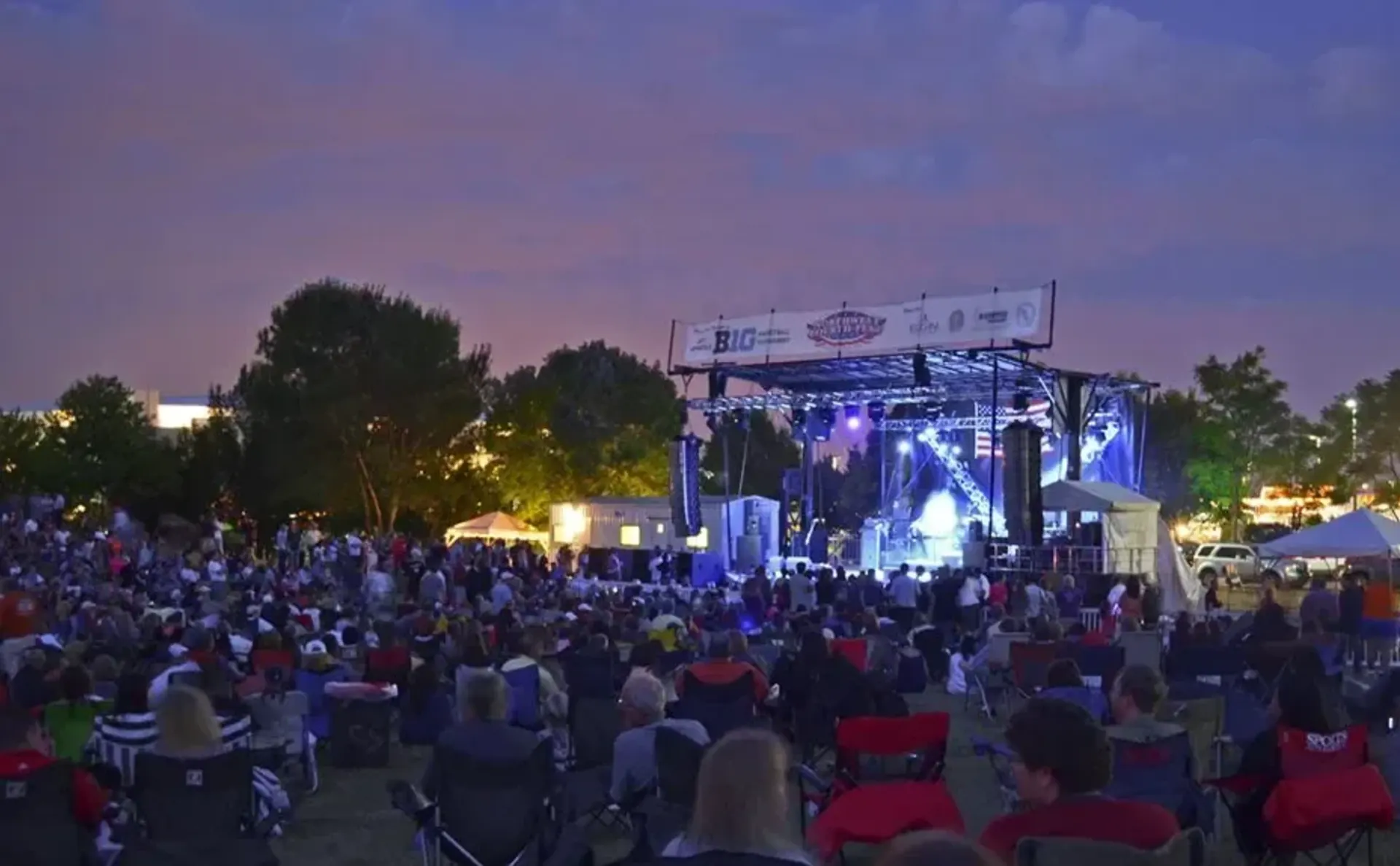 Community concert series at dusk with large lawn crowd and truss roof stage with concert lighting