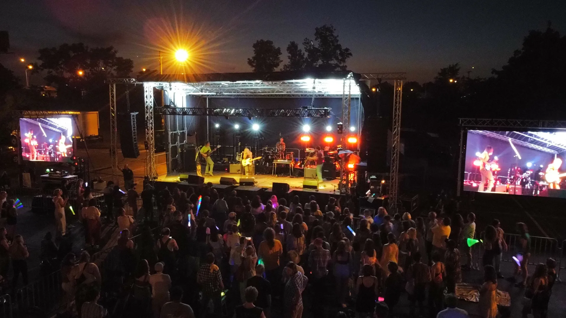 Outdoor evening concert with LED IMAG screen flanking stage and crowd with glow sticks