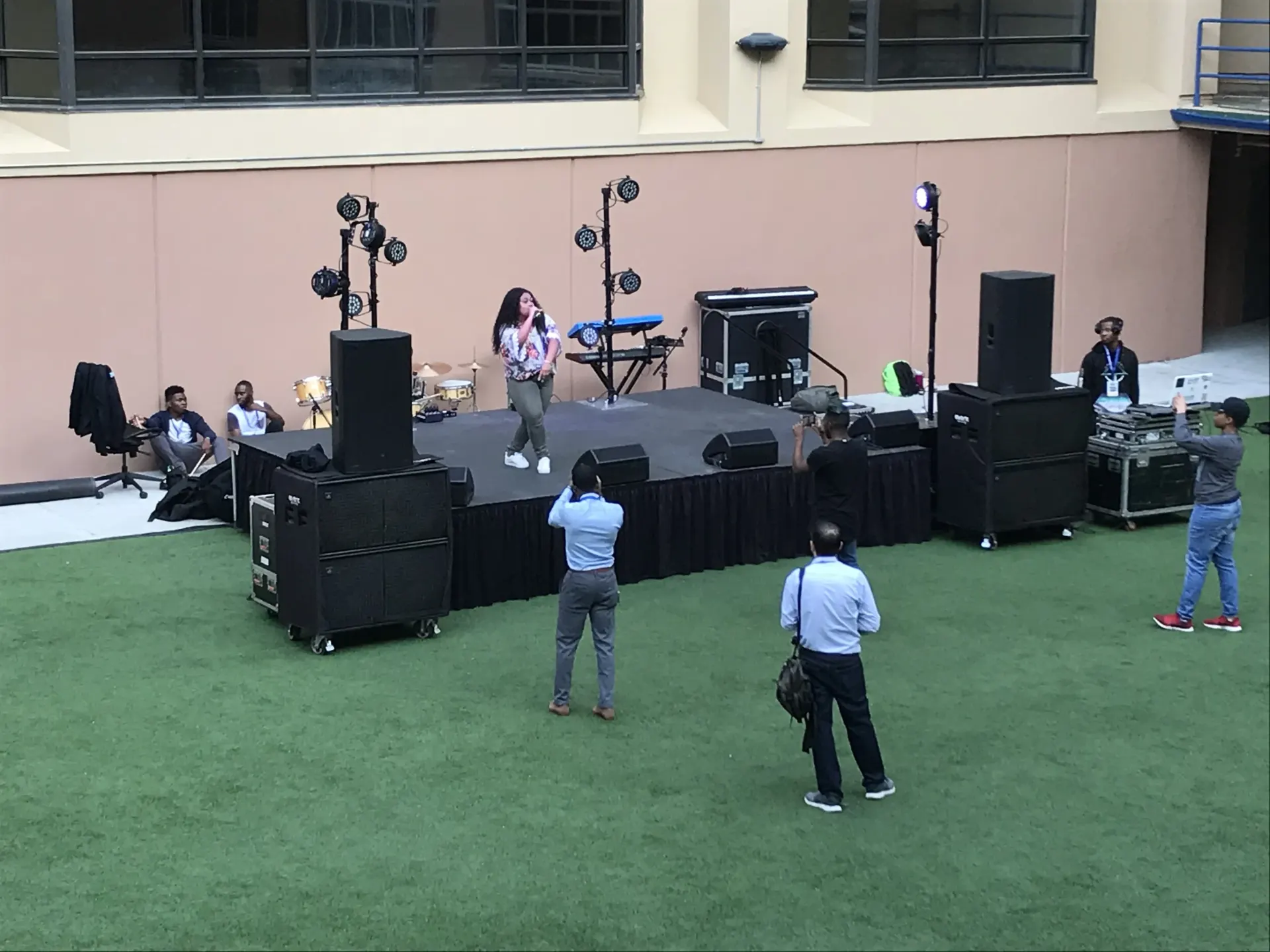 Singer performing on skirted deck stage with lighting and PA speakers in outdoor courtyard