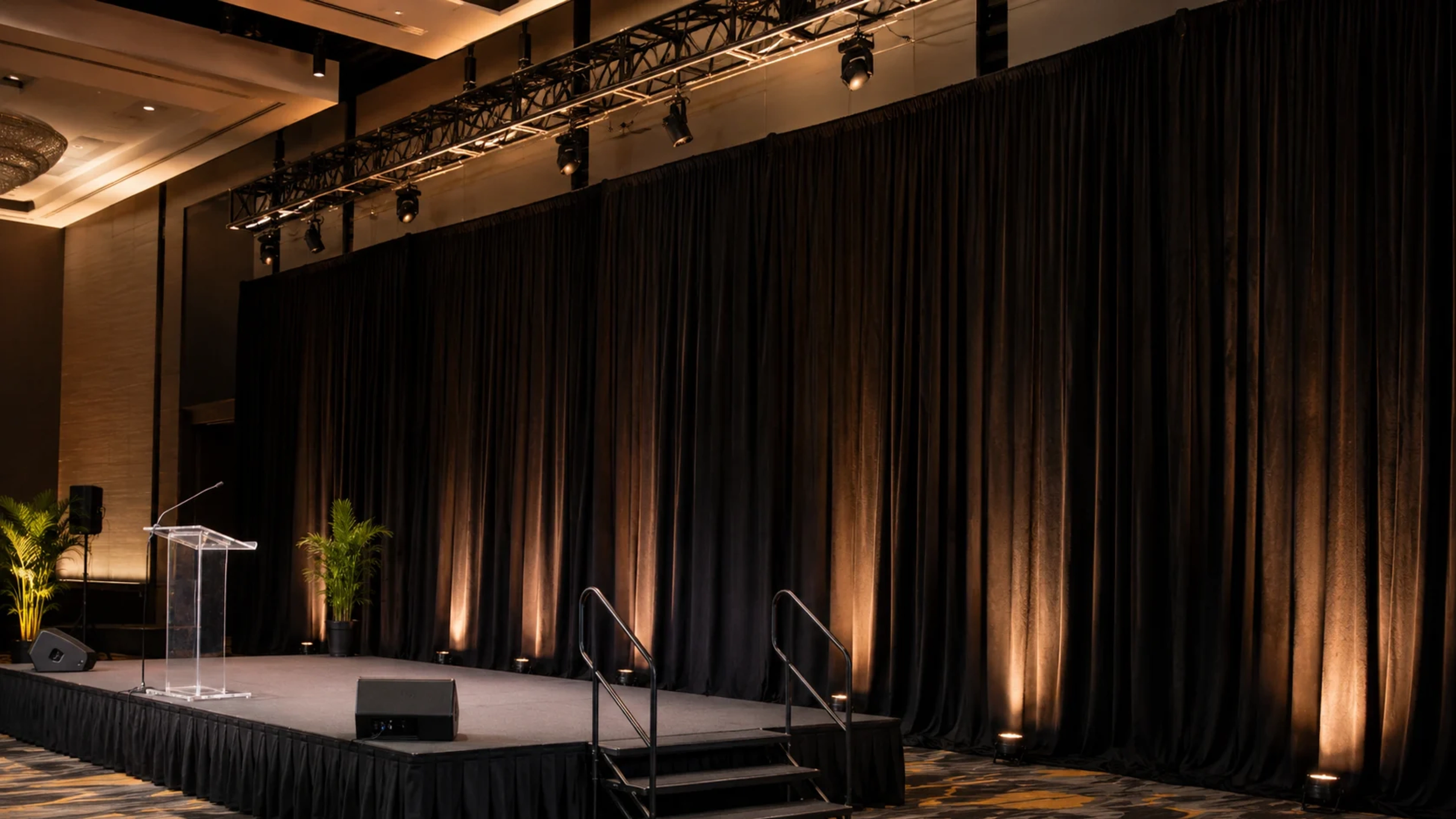 Full black pipe and drape backdrop behind a ballroom stage with warm uplighting, a clear lectern, and overhead truss