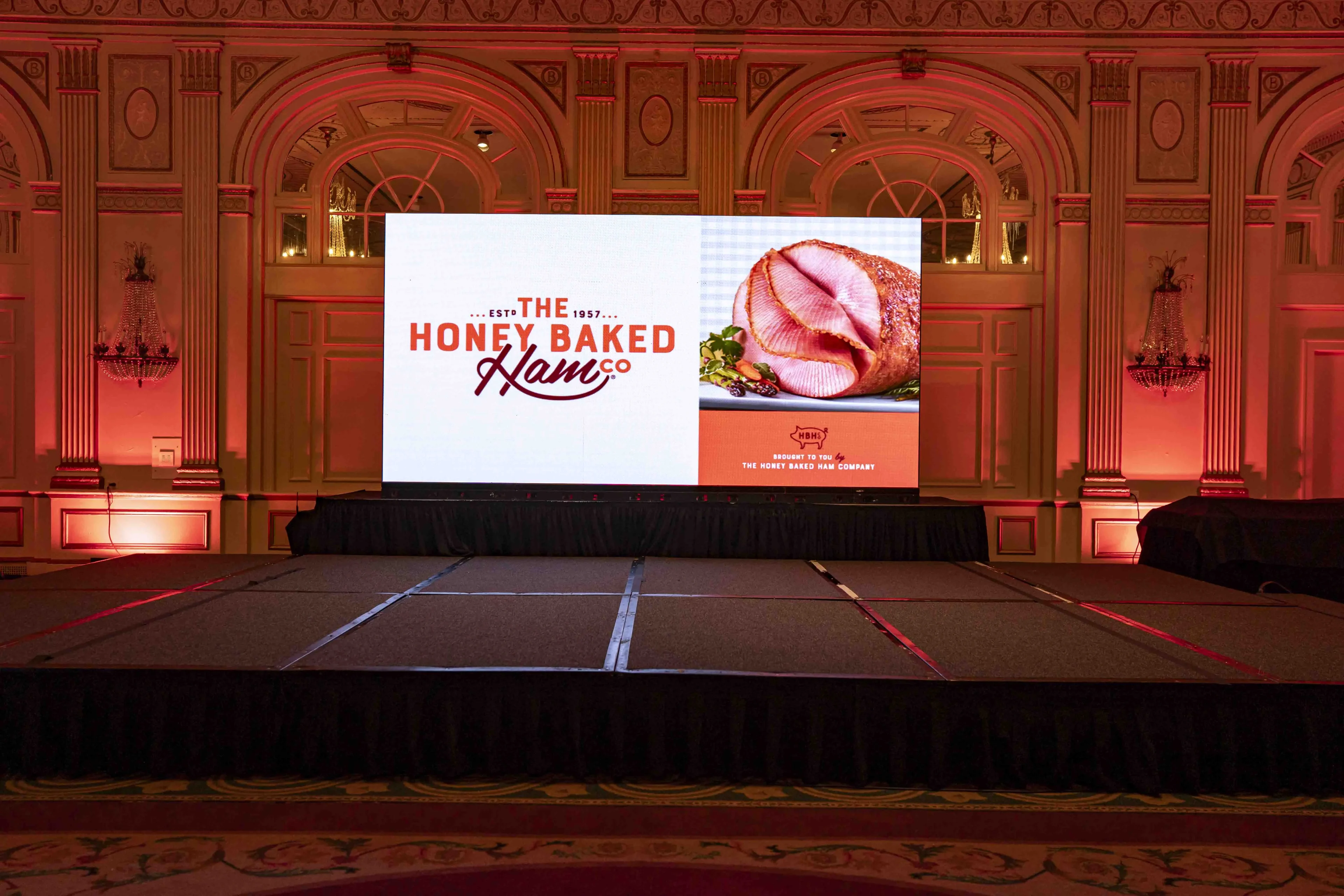 HoneyBaked Ham brand-reveal LED screen with 'Est 1957' logo and product imagery against a red-washed historic ballroom
