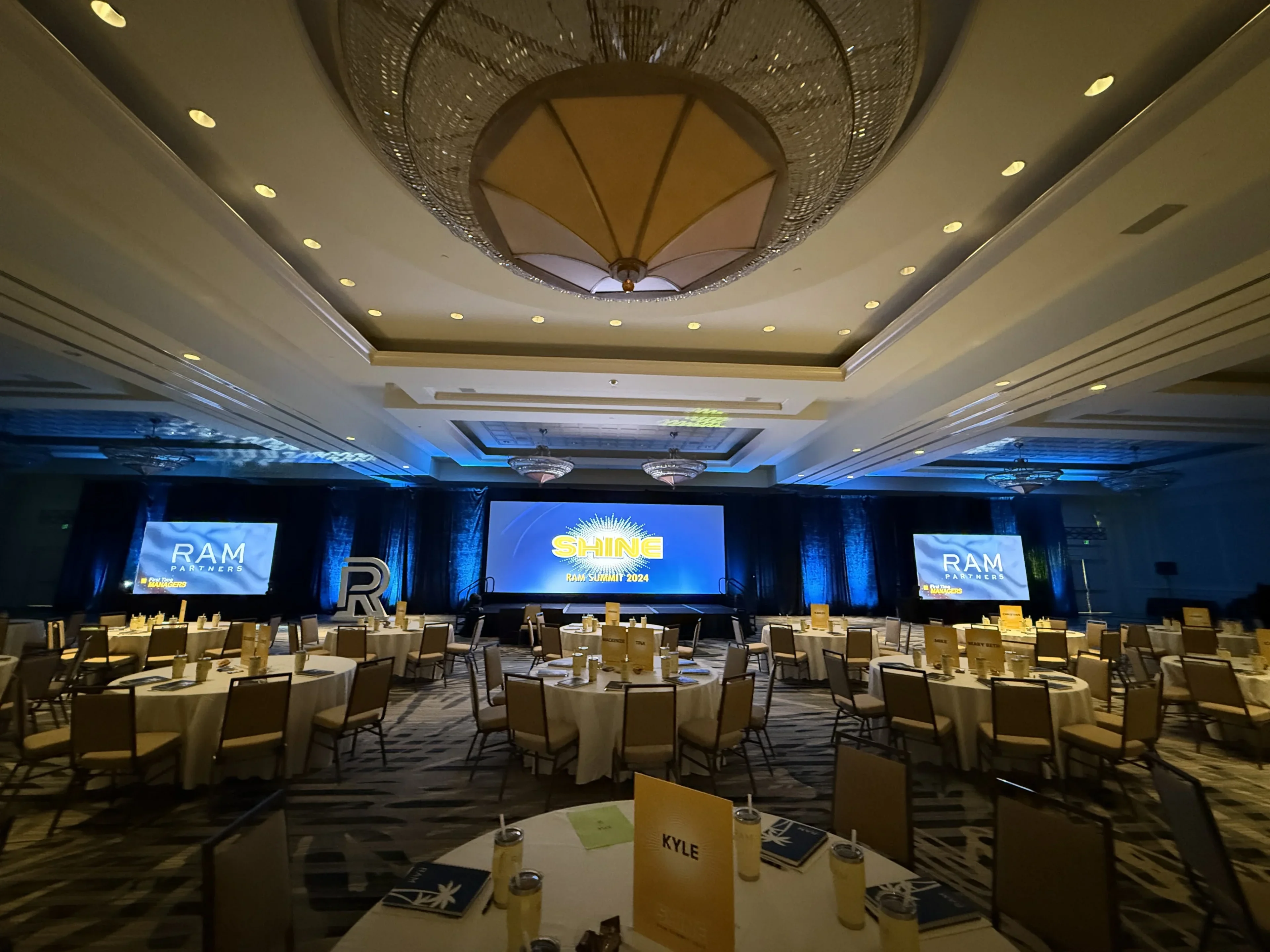 RAM Partners Shine 2024 Summit ballroom with center LED wall and twin IMAG screens, banquet rounds, and blue wash lighting
