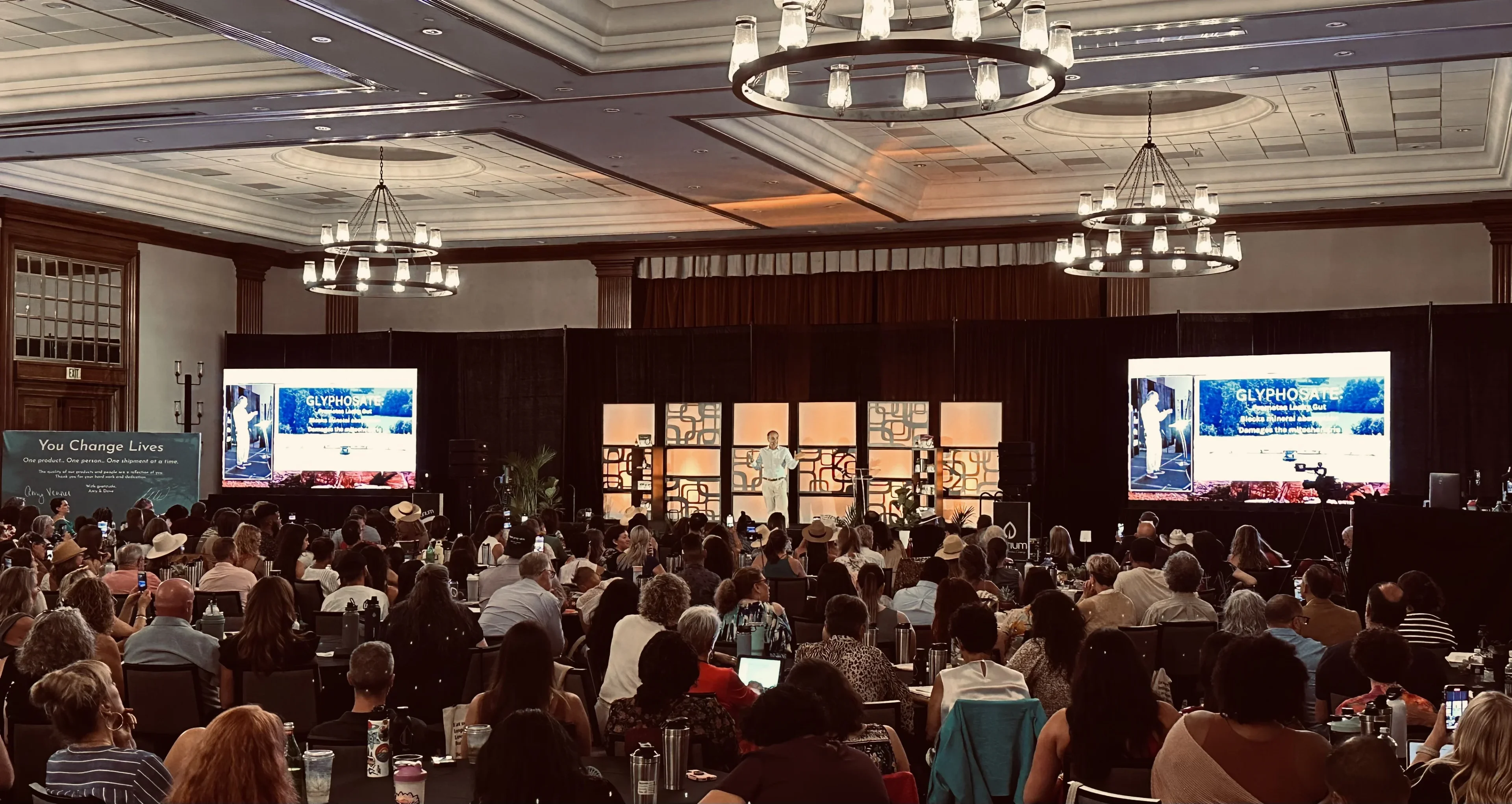 Purium general session in a wood-paneled ballroom with dual side LED screens showing the keynote speaker and a packed audience at rounds