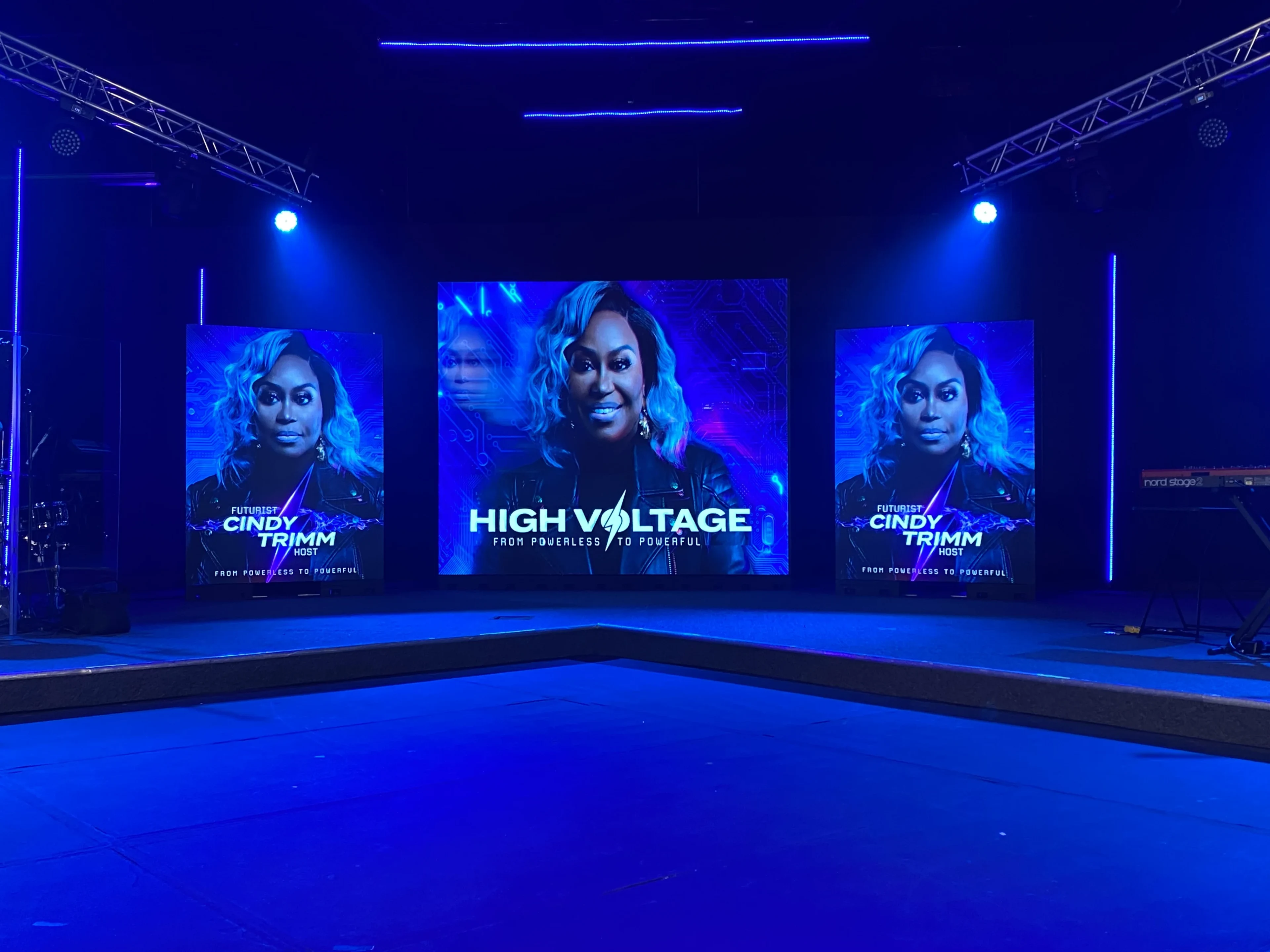 High Voltage 'From Powerless to Powerful' stage with three LED panels — Cindy Trimm host portraits flanking a center title panel — under truss with movers and a deep blue wash