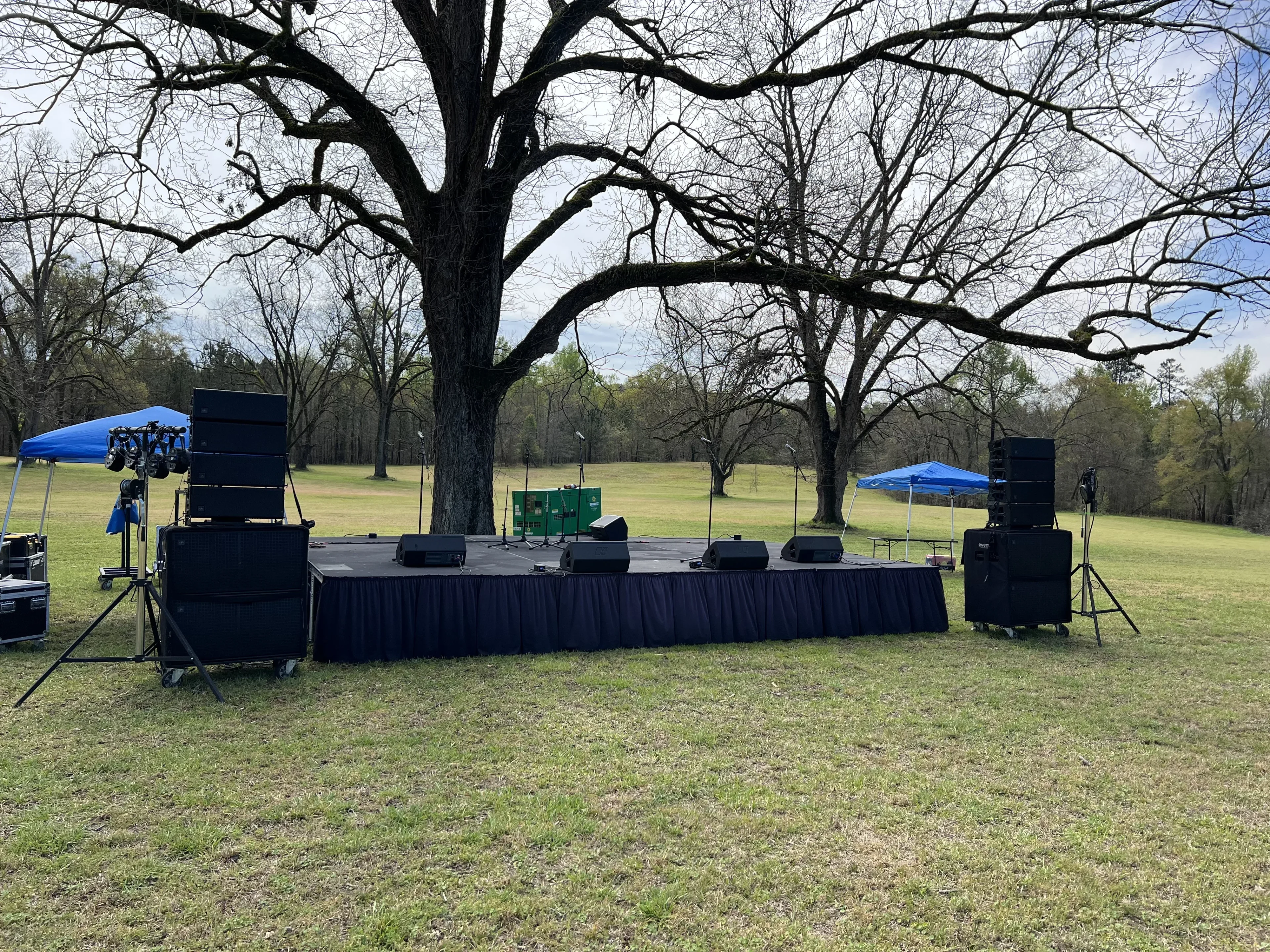 Skirted deck stage with PA speakers and monitors set up in a park field for outdoor event