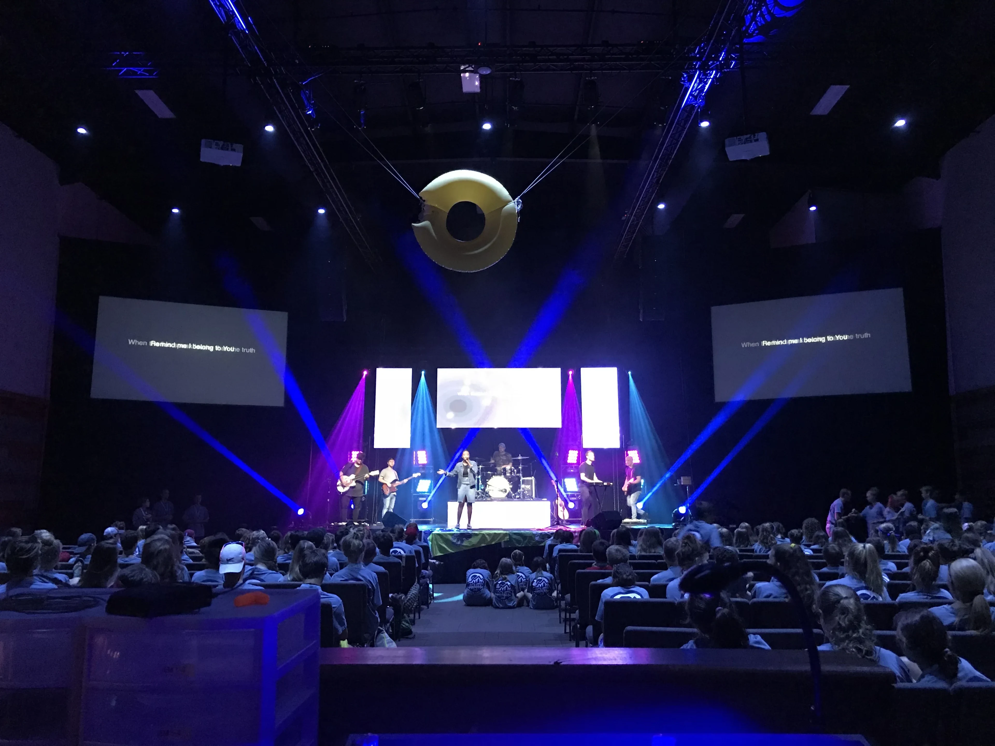 Youth worship service with full band, blue beam lighting, LED screens, and engaged audience in church auditorium