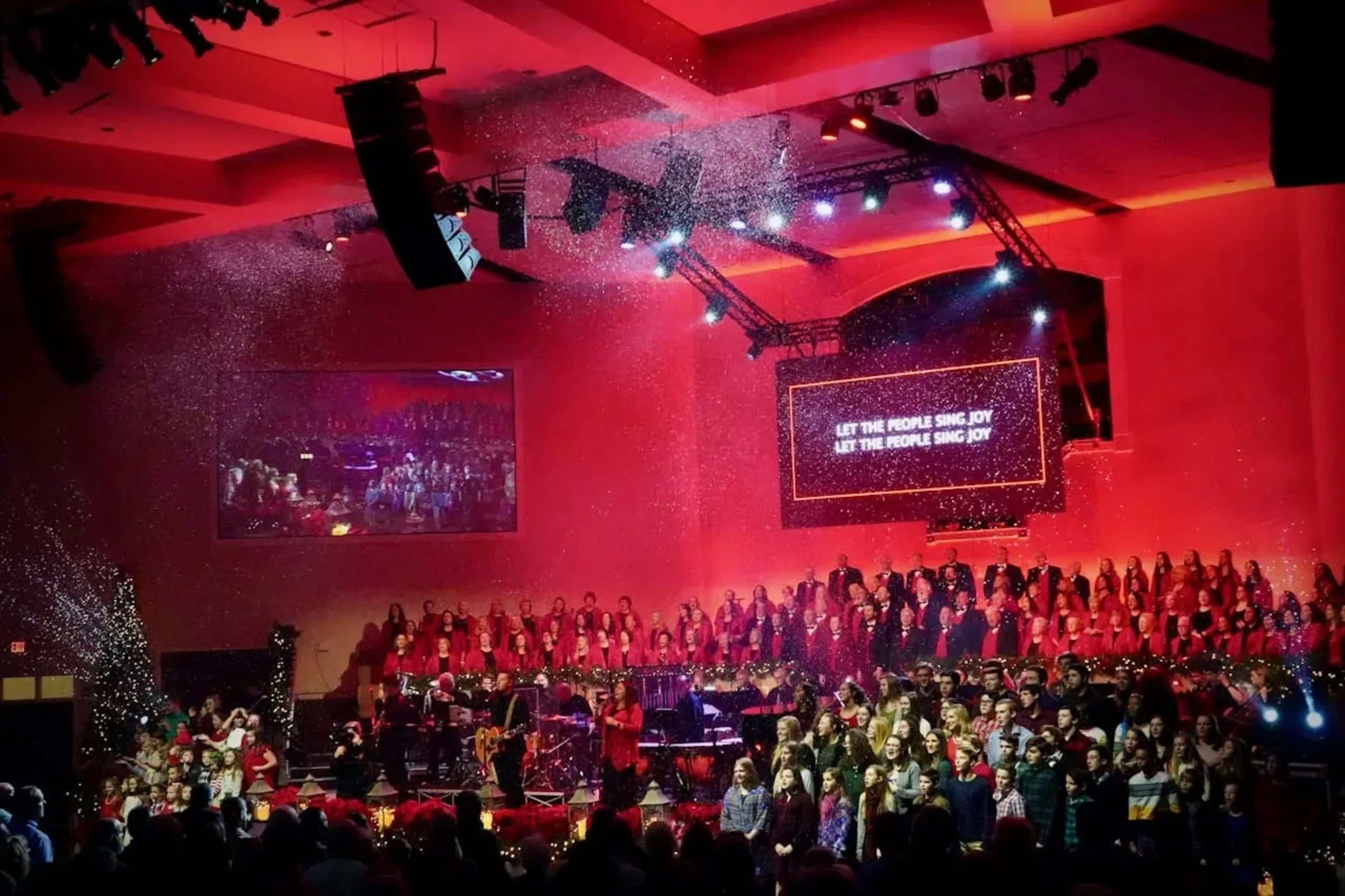 Large church Christmas celebration with full choir, red stage lighting, snow effects, and LED screens