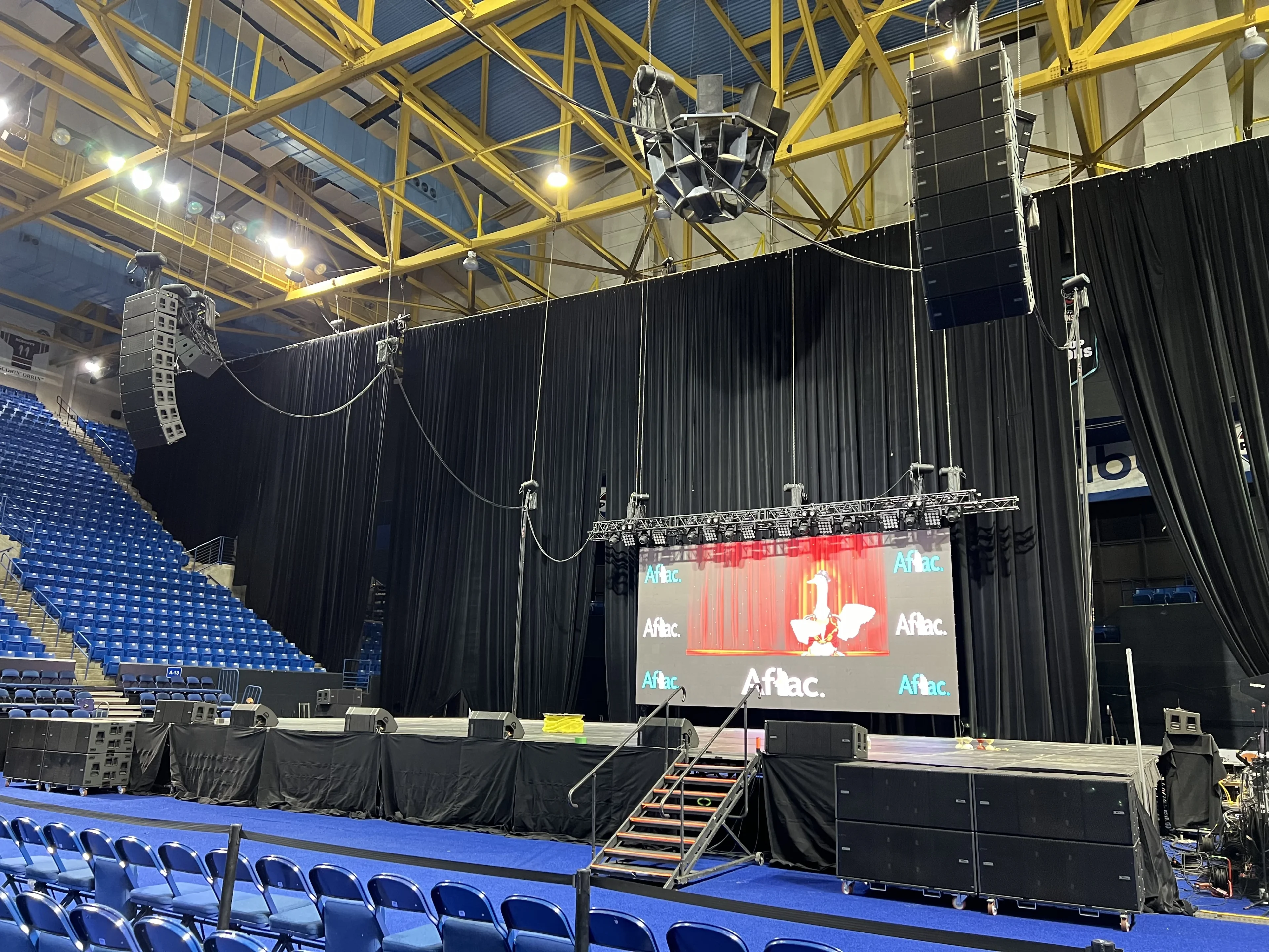 Indoor arena with ceiling-rigged truss grid, visible chain motors, LED video wall on stage, and flown line array speakers