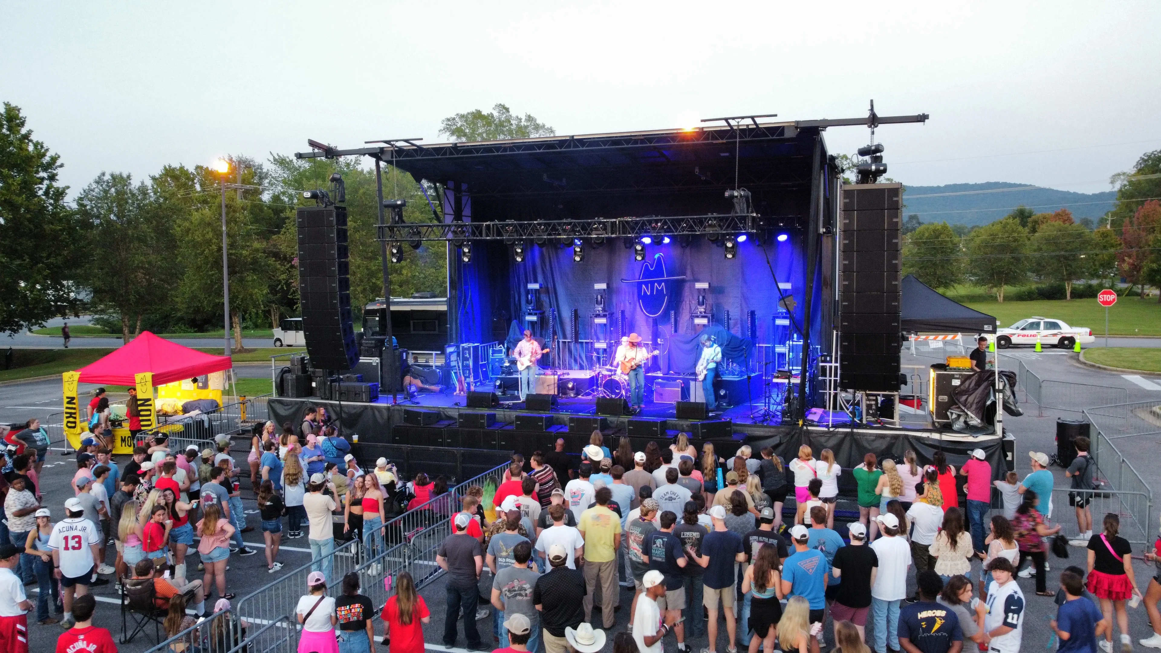 Live outdoor concert with truss roof system supporting mounted lighting fixtures and flown PA line arrays over performing band