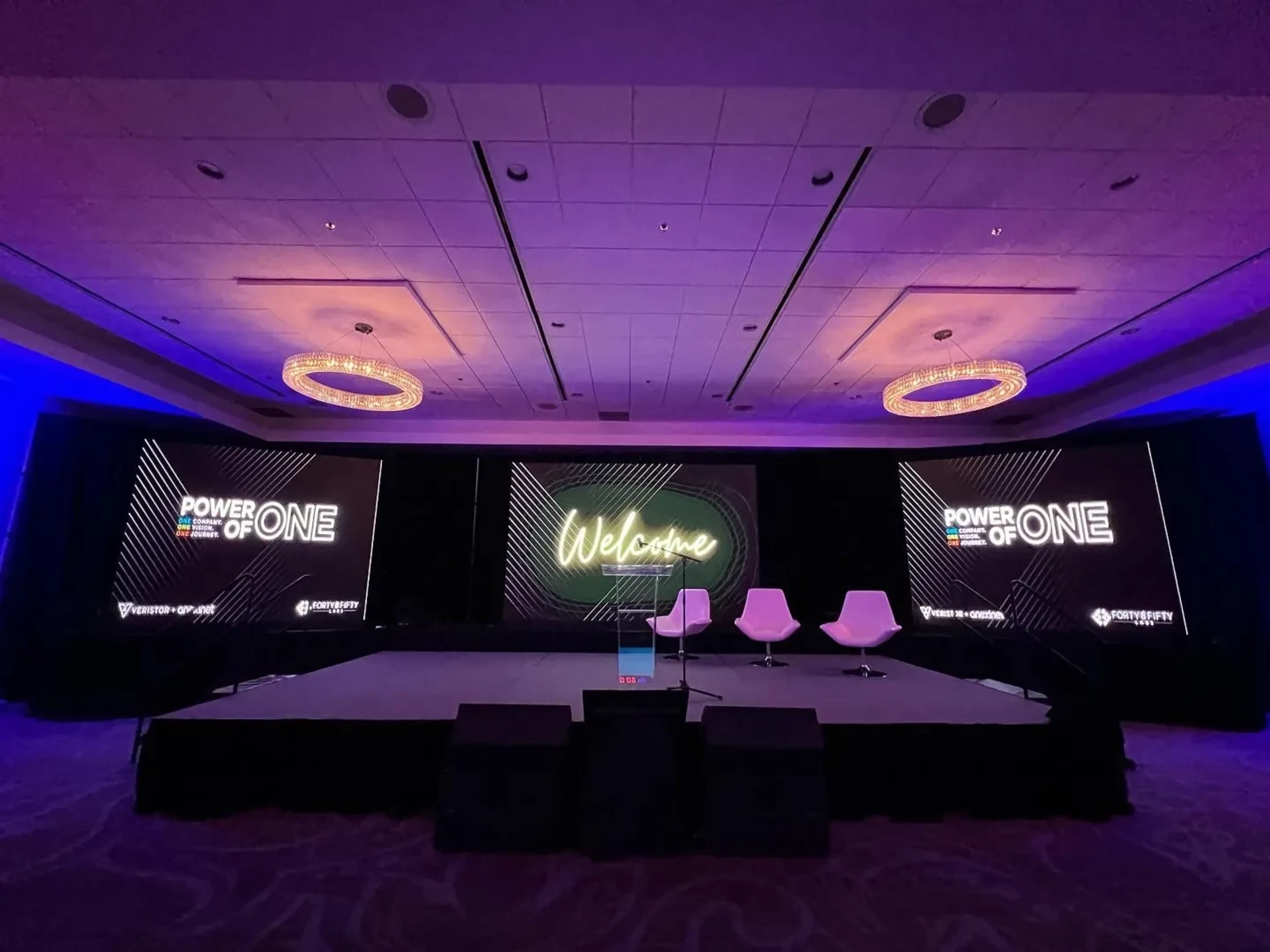 Power of One conference stage with three LED panels — branded side panels and a 'Welcome' center graphic — over a deck stage with three lounge chairs and purple uplighting