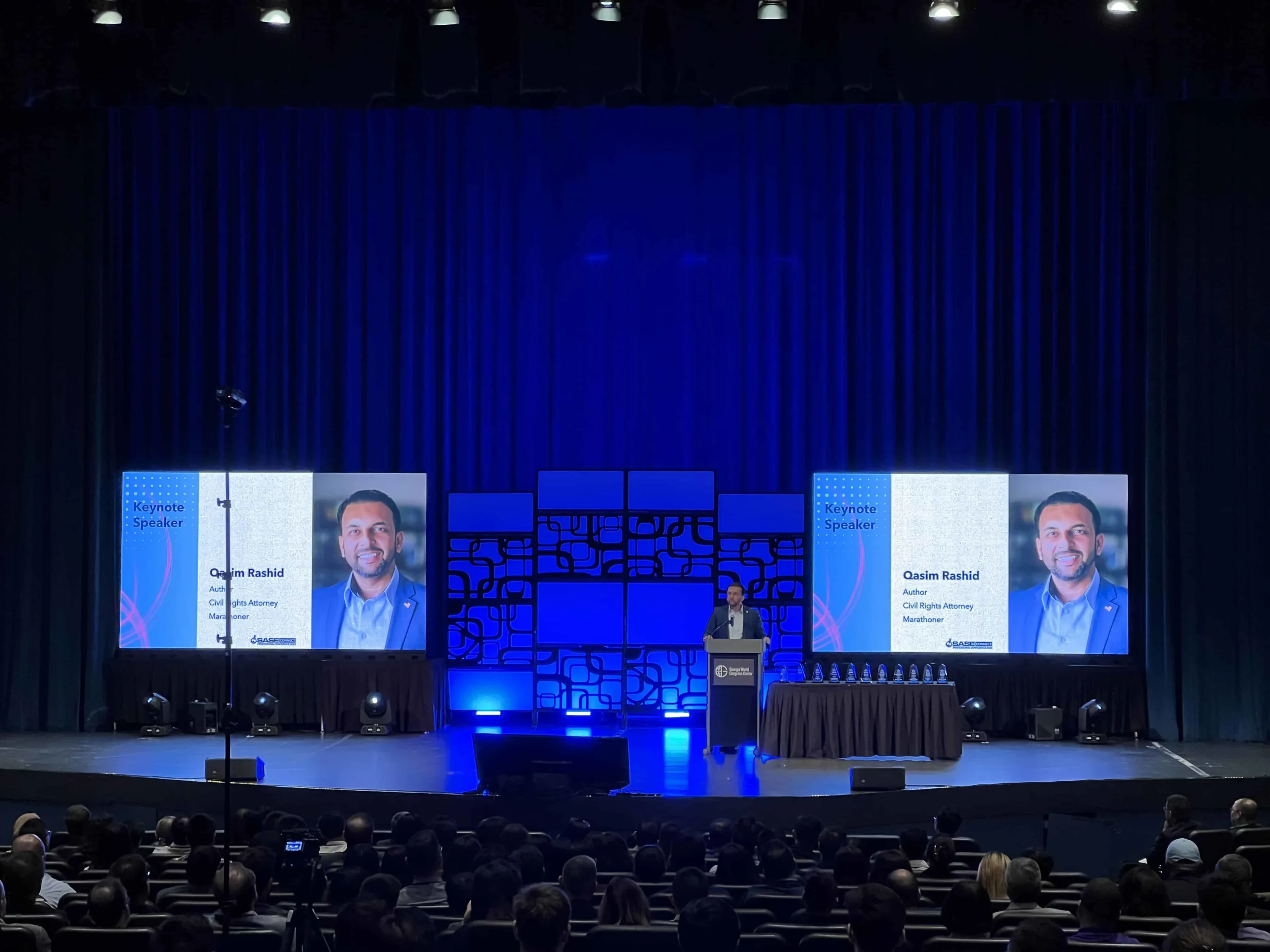 SASE Summit keynote in progress with Qasim Rashid at the lectern, dual large IMAG LED screens showing his speaker graphic, a custom geometric stage backdrop illuminated in blue, and a full audience in silhouette
